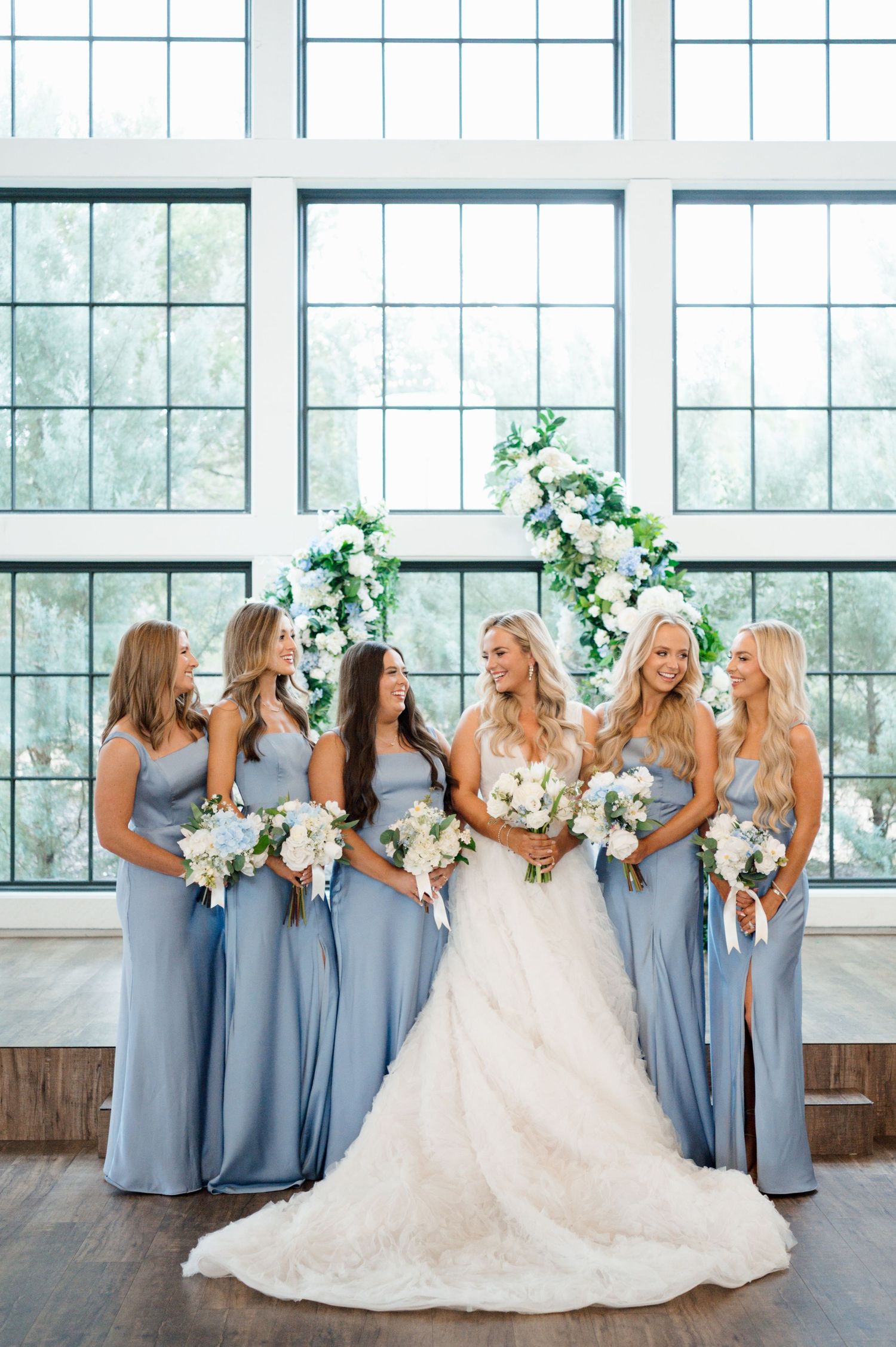 A bride in a white wedding dress is standing with her bridesmaids, who are wearing gray dresses, in front of large windows.