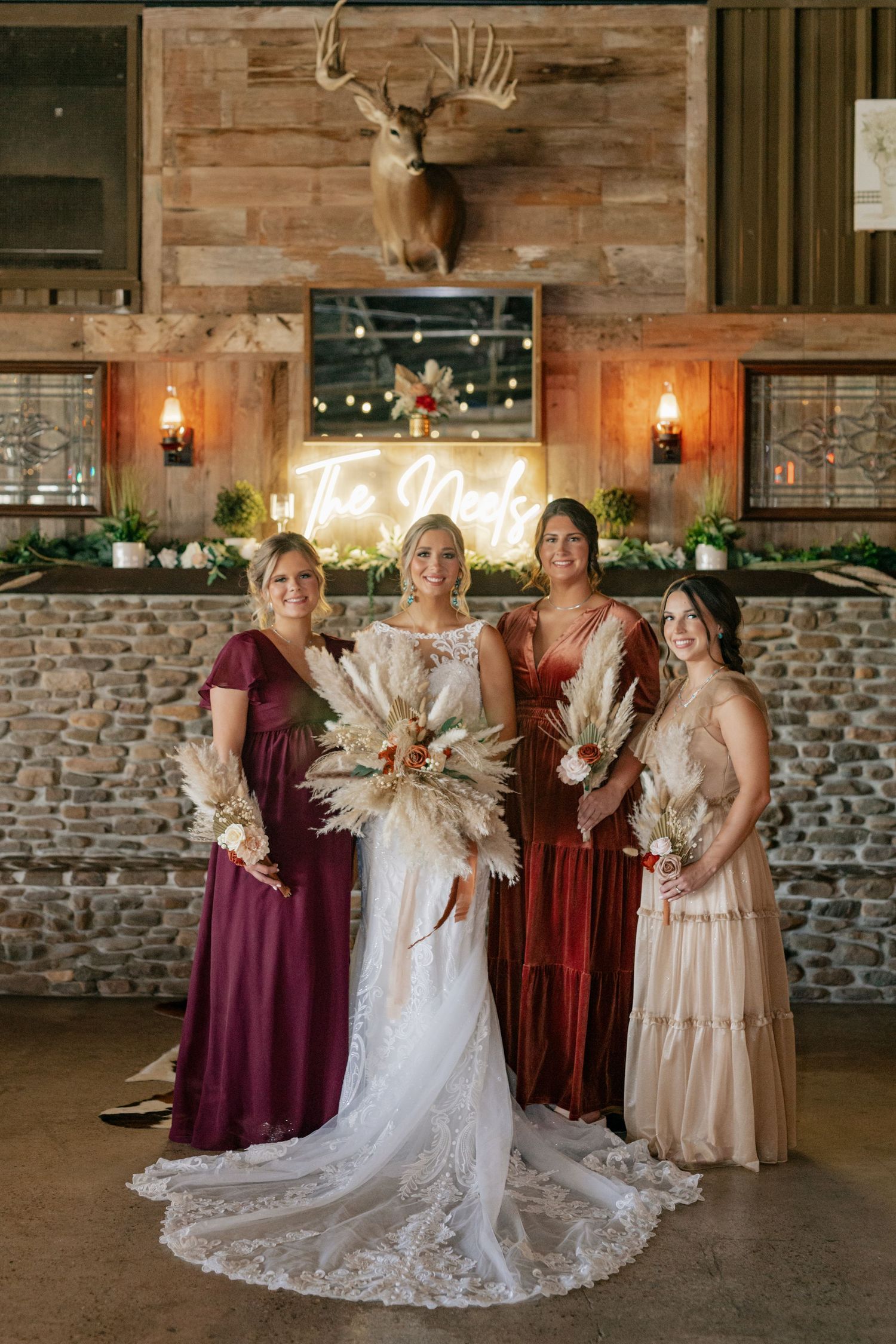 Wedding party in burgundy and blush dresses posing under mounted deer head and neon sign.