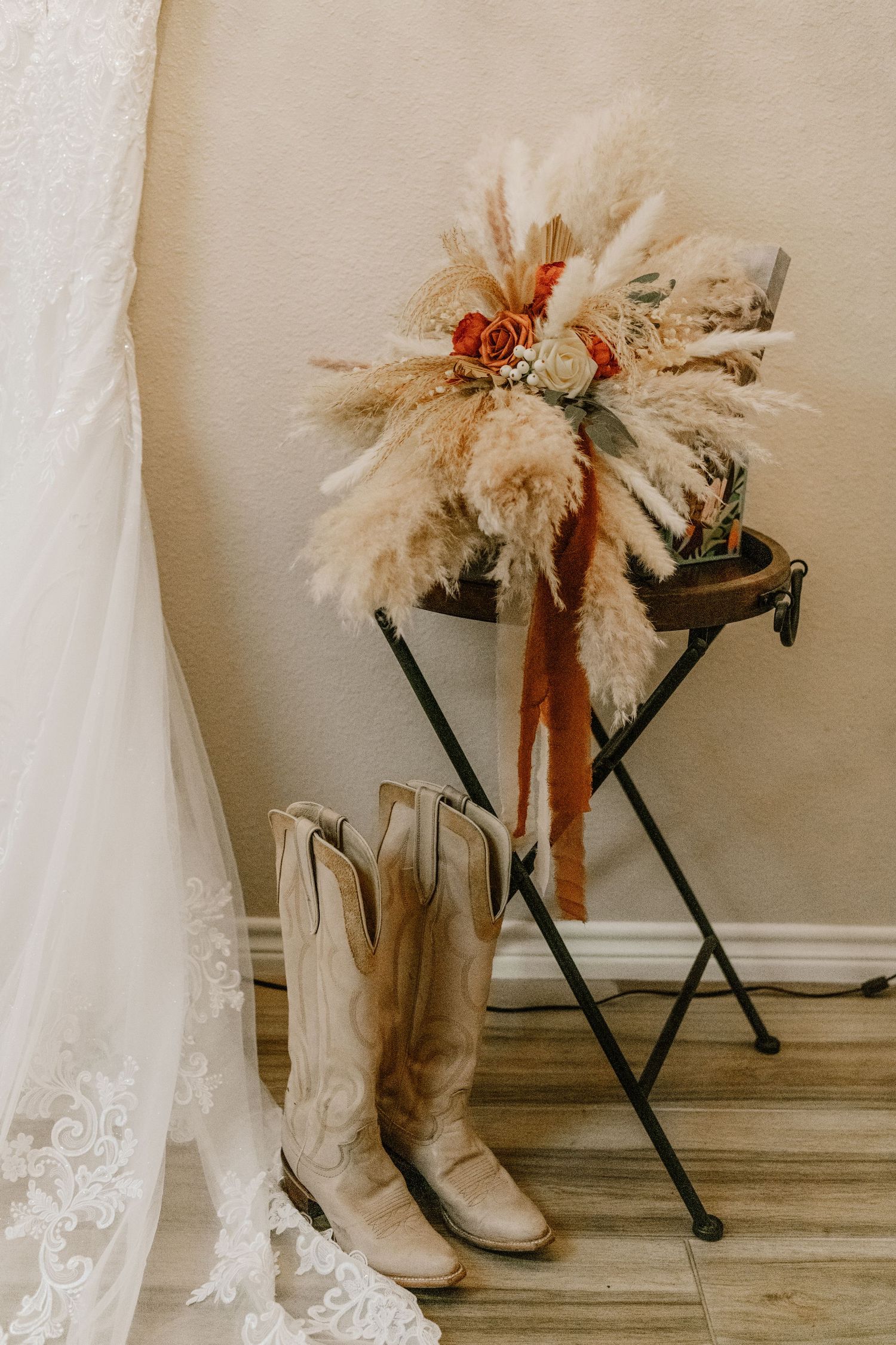 White cowboy boots and a dried pampas grass floral arrangement against a wall with lace wedding dress fabric.