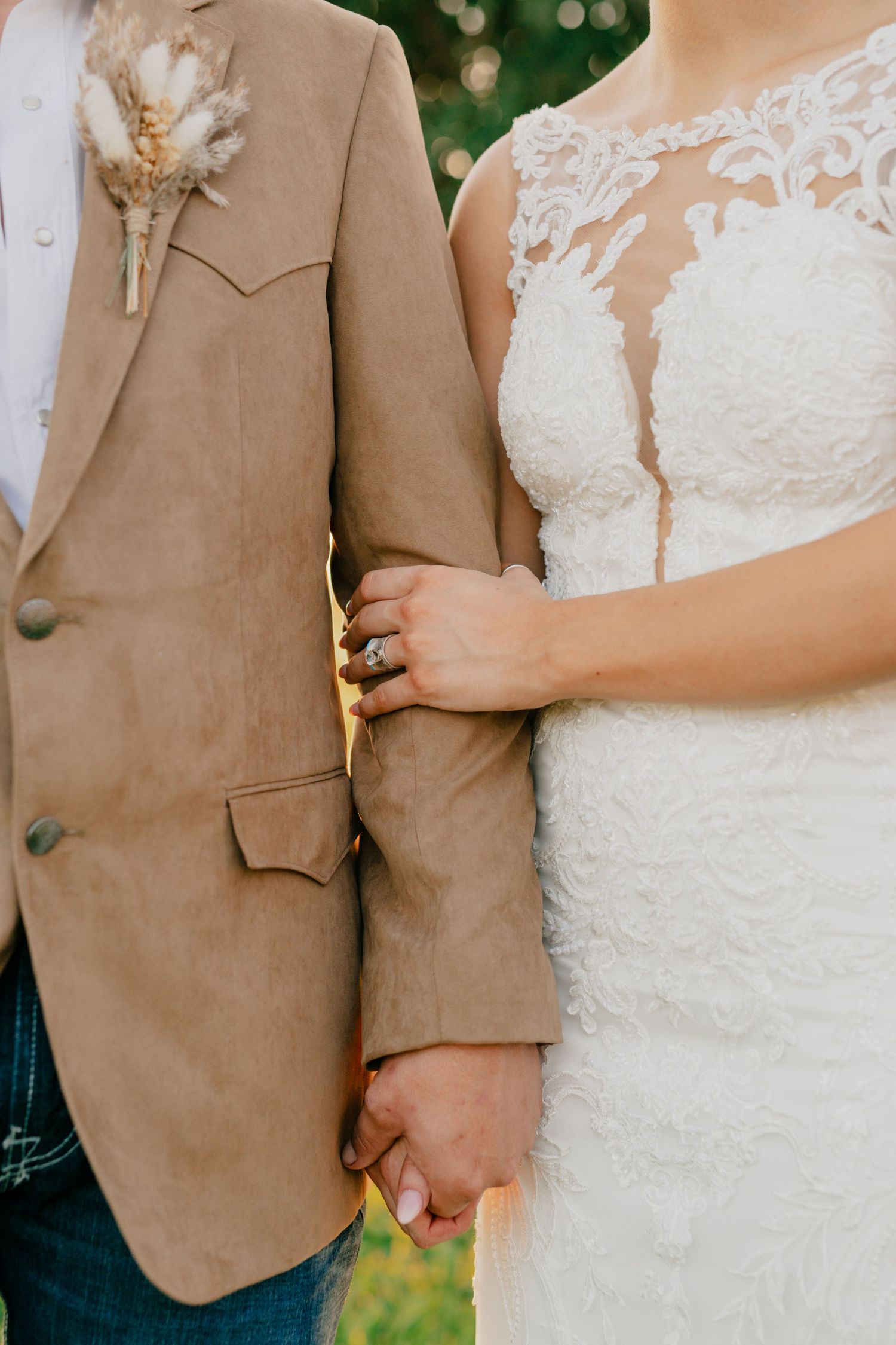 Close-up view of a couple holding hands at their wedding, showing a tan suit jacket and white lace dress details.