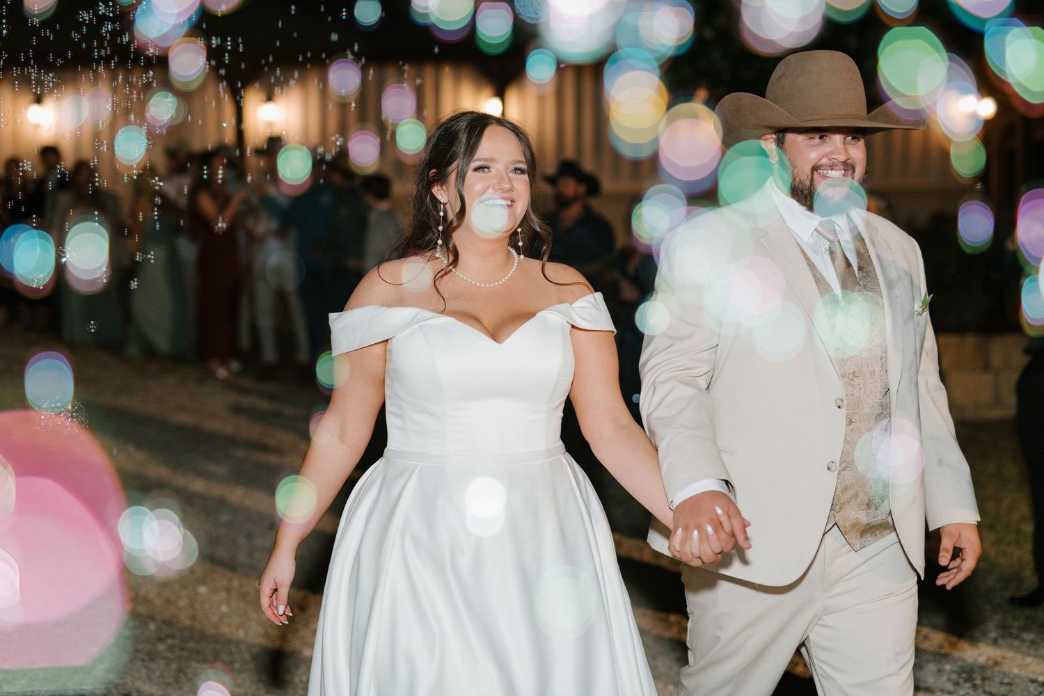 A couple walks together at an evening wedding celebration under twinkling string lights and bokeh effects.