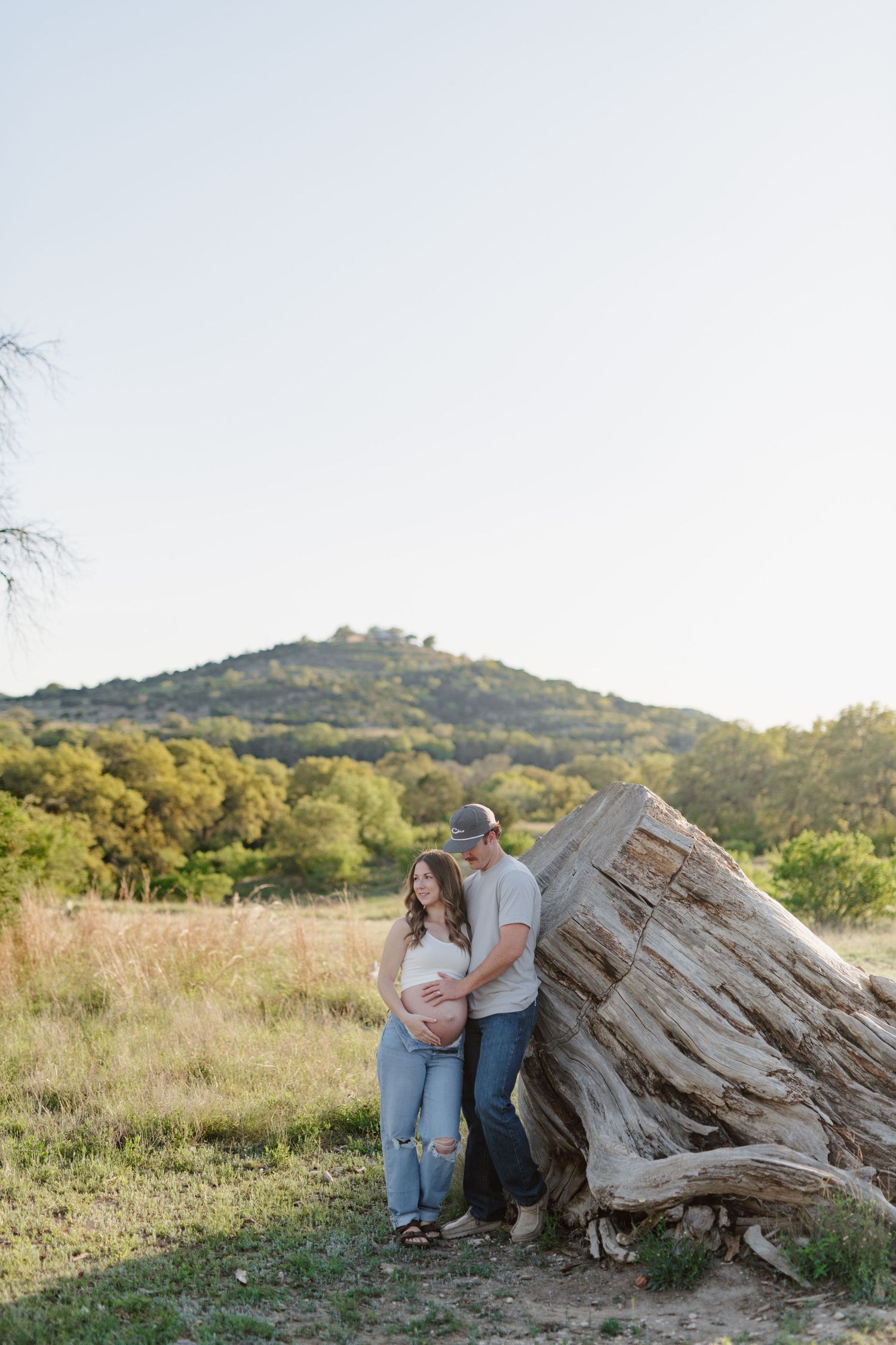 Couple stands together near large driftwood in scenic hill country field during golden hour sunset.
