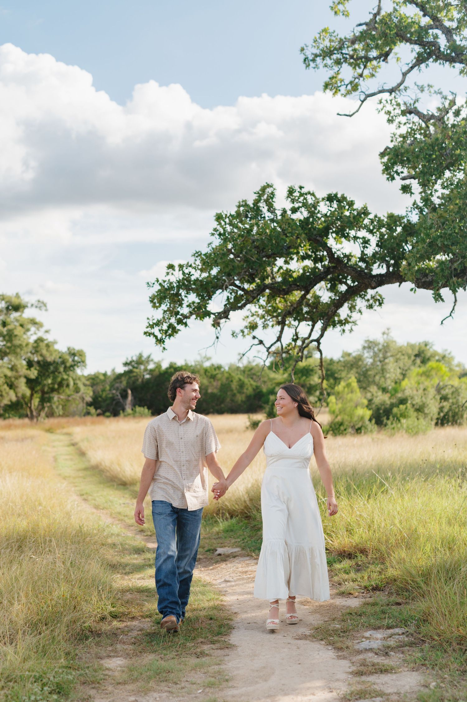 A couple walking hand in hand along a winding dirt path through a sunny meadow.