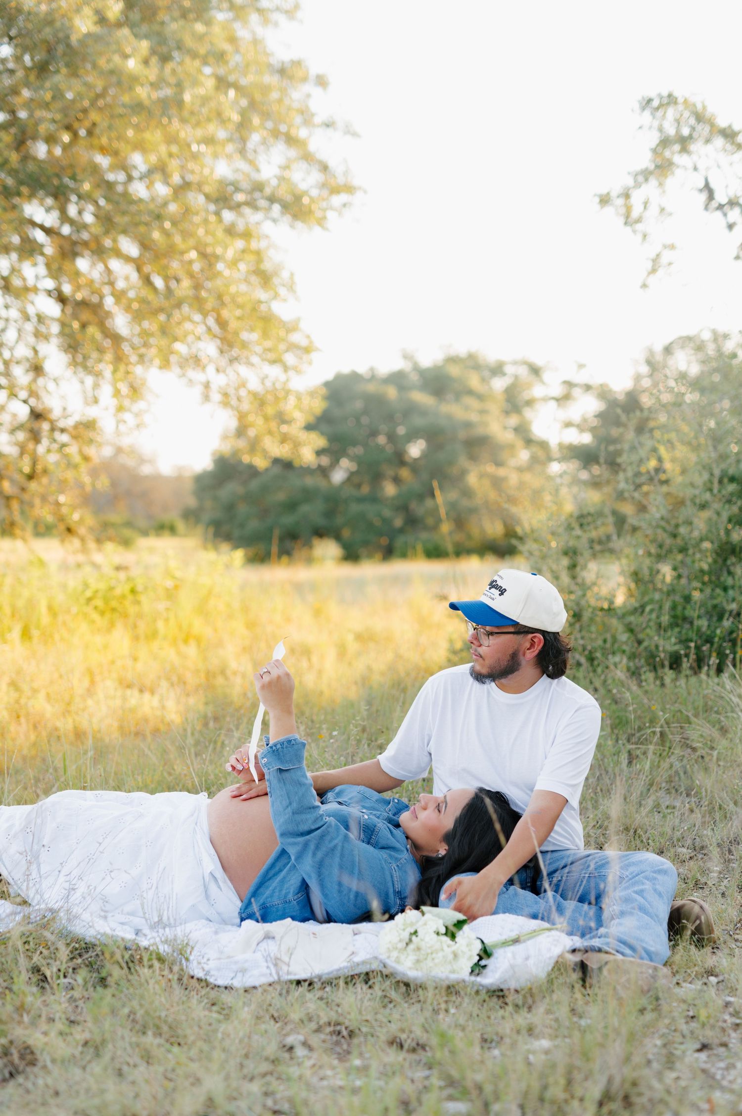 A couple relaxes on a white blanket in a golden sunlit field during their maternity photo session.