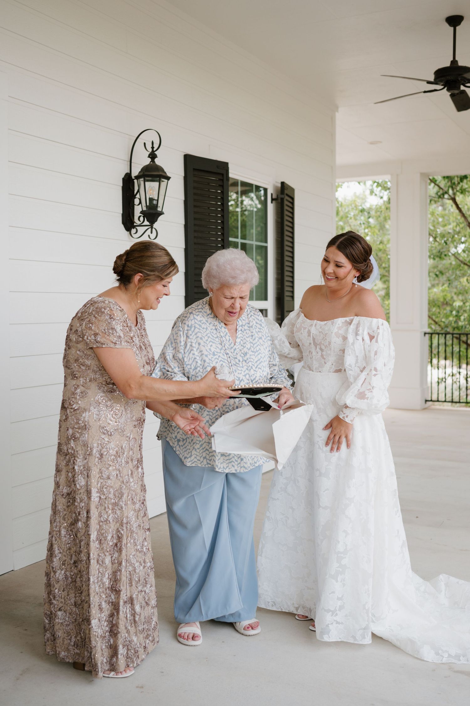 Three women share an emotional moment while looking at photos on a sunny white porch.