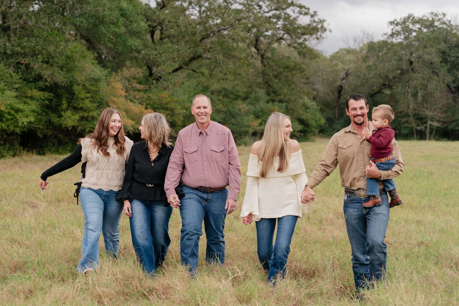 A family walks together holding hands in a grassy field with oak trees in the background.