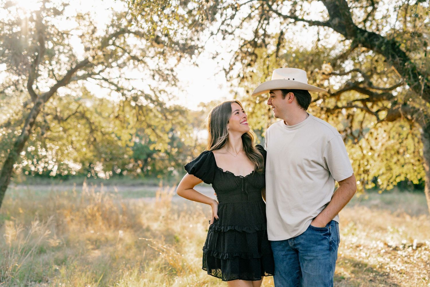 A couple laughs together while posing in casual attire against a backdrop of sunlit trees.