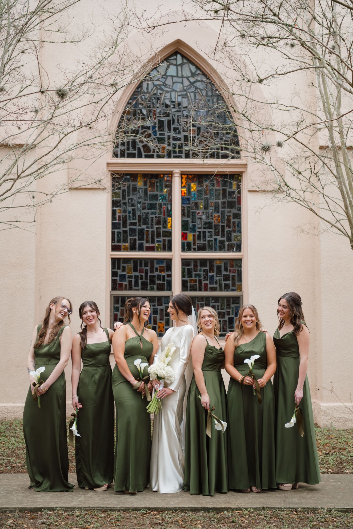 A wedding party poses in front of a church window wearing elegant olive green bridesmaid dresses and a white bridal gown.