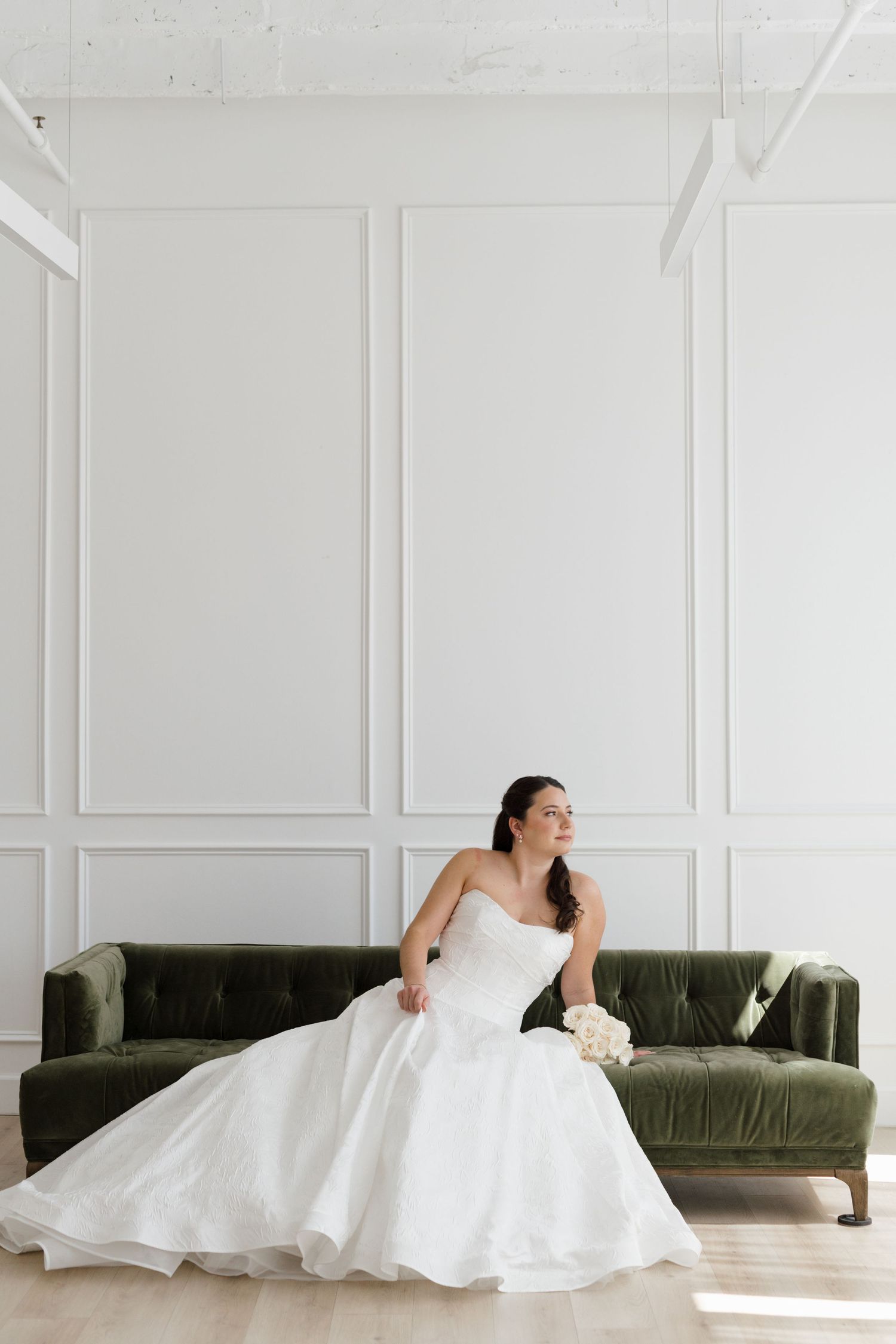 Bride in strapless white ball gown sits on green velvet sofa holding bouquet in bright white paneled room.