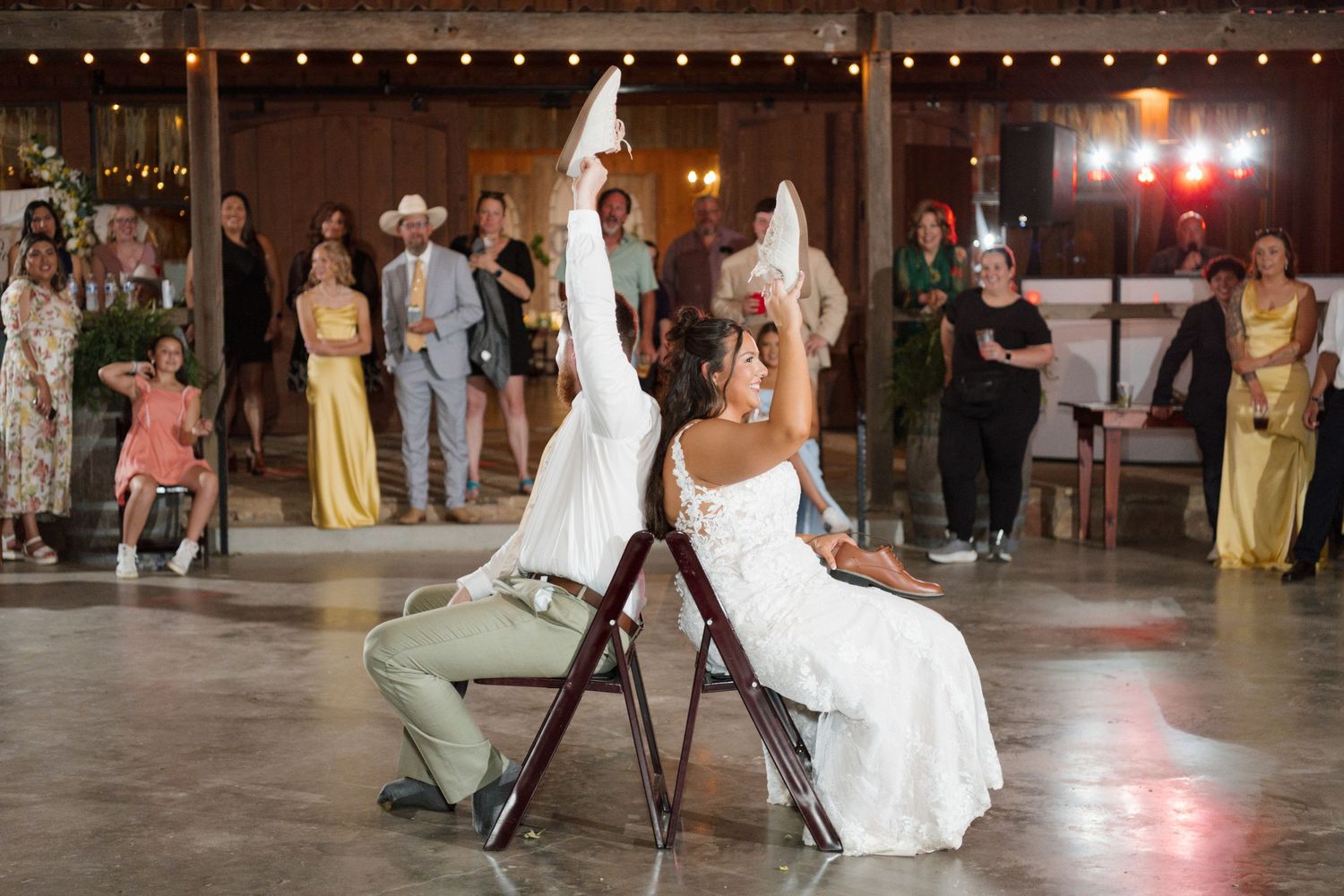 Groom playfully removes bride's garter as she raises her leg during a fun barn wedding reception with cheering guests.