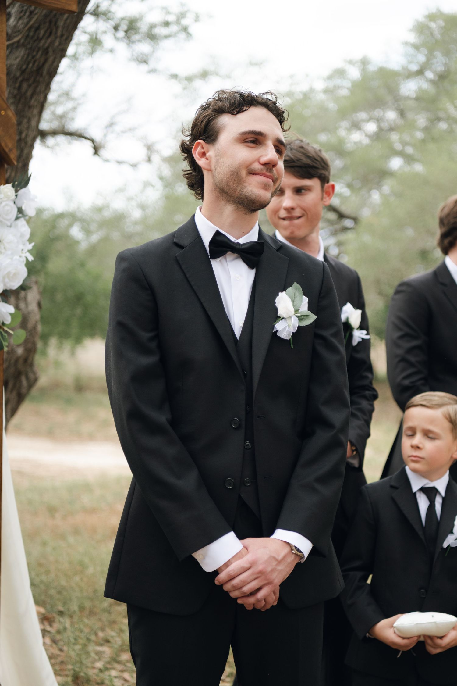 Groom in black tuxedo with white boutonniere stands at altar during outdoor wedding ceremony.