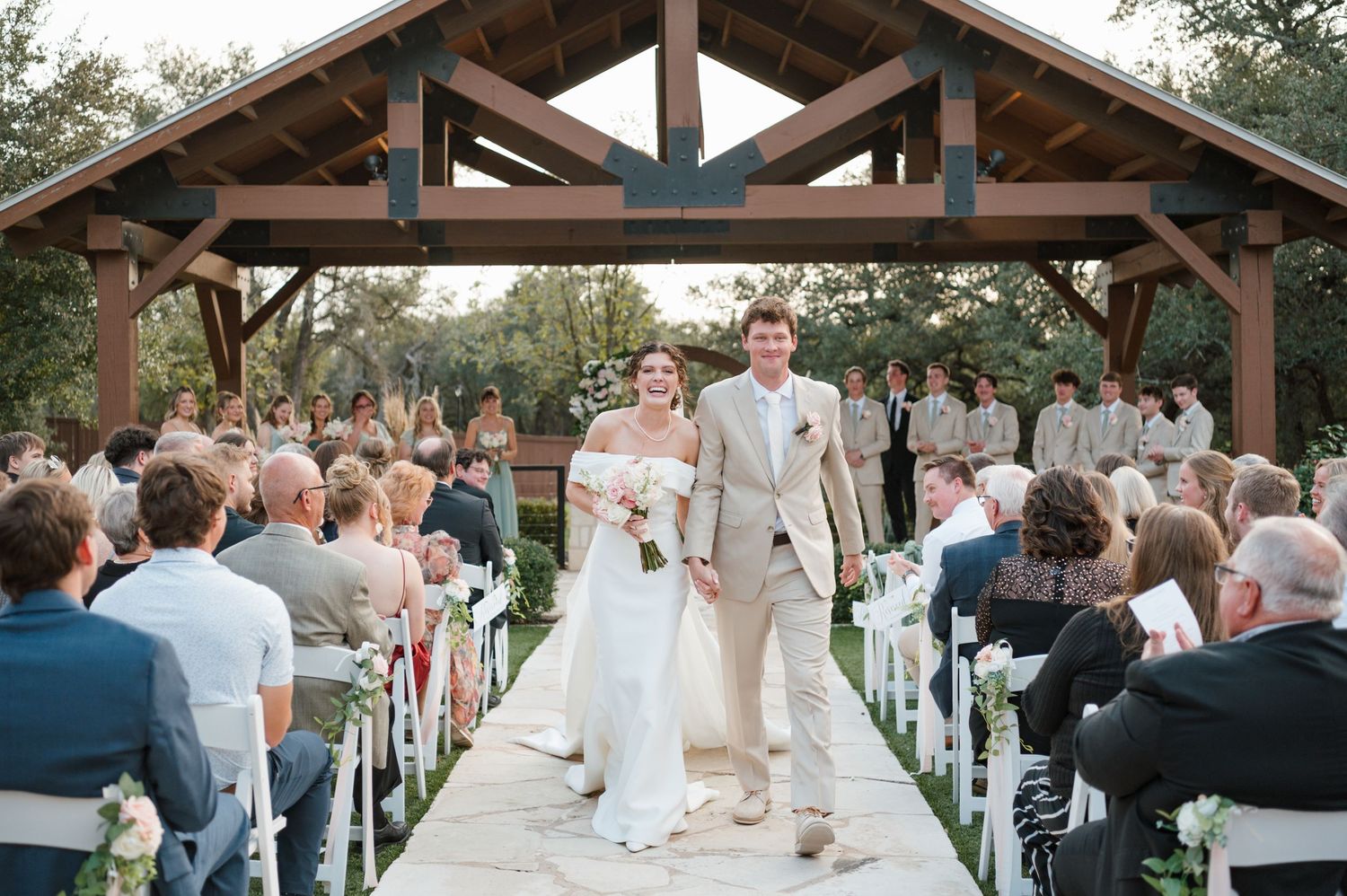 Outdoor wedding ceremony under rustic wooden pavilion with guests seated in white chairs and natural greenery surroundings.