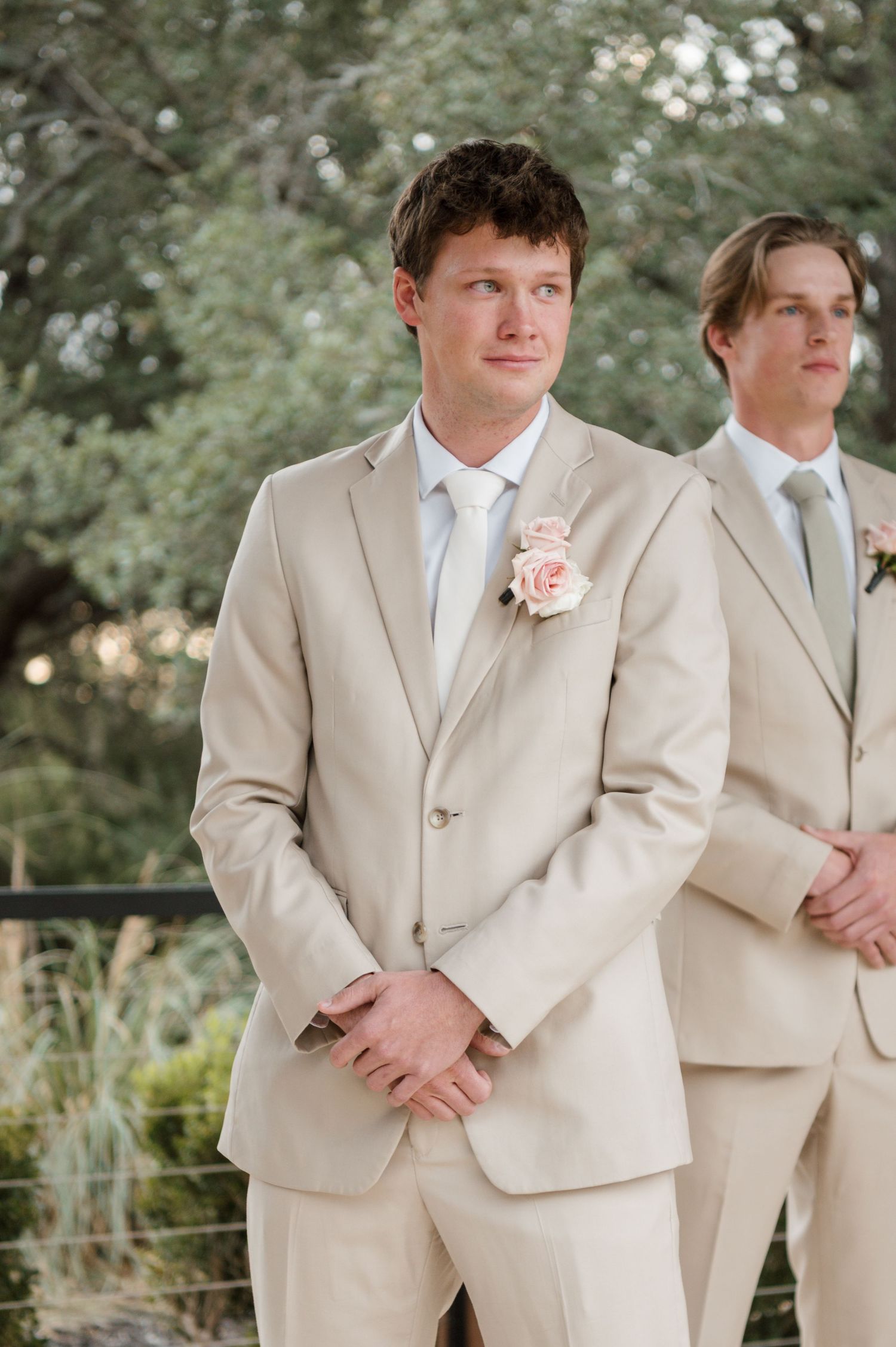 Groomsmen in beige suits stand outdoors during wedding ceremony with natural backdrop.
