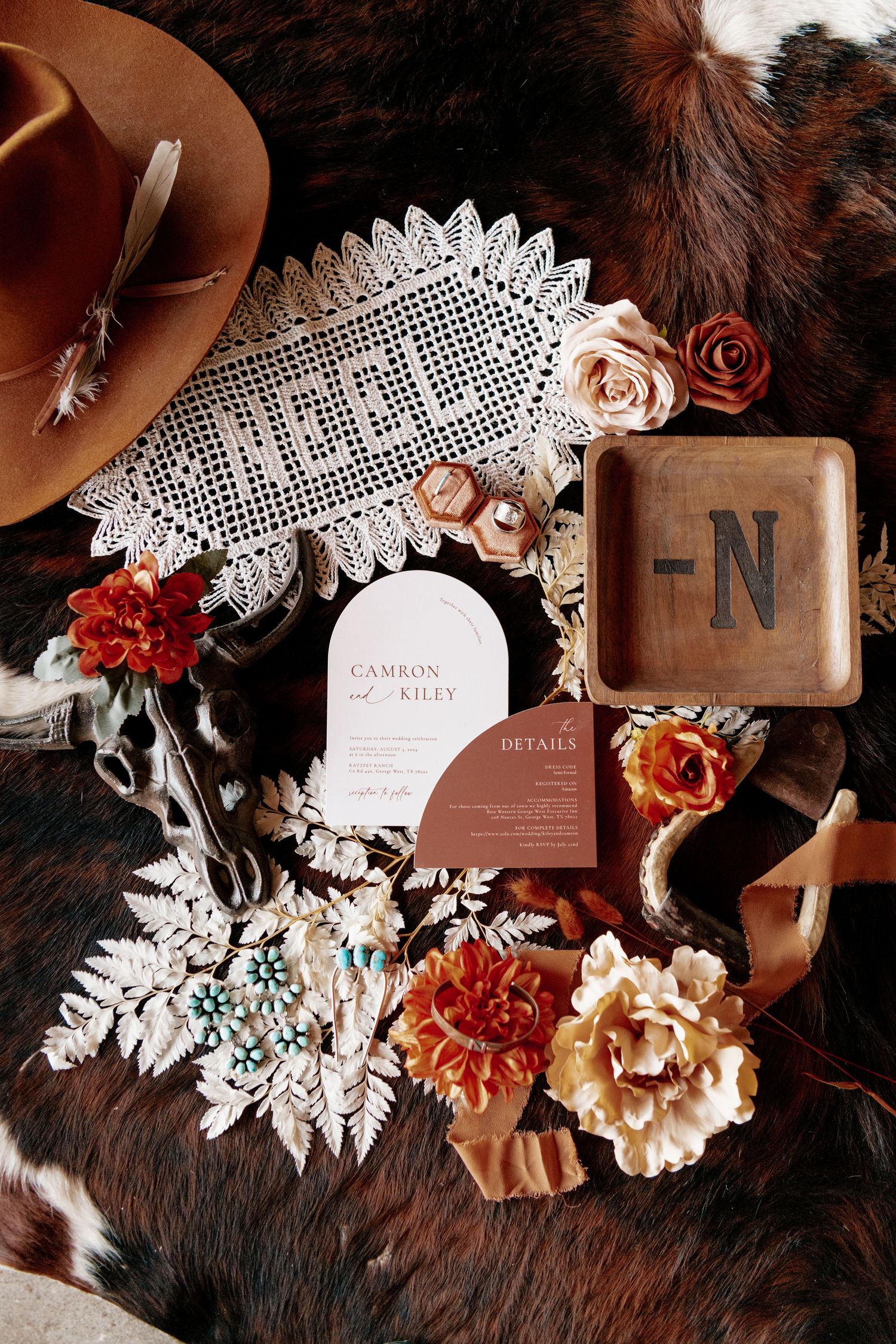 Rustic wedding details featuring a brown hat, lace doily, floral arrangements and invitation cards on cowhide.