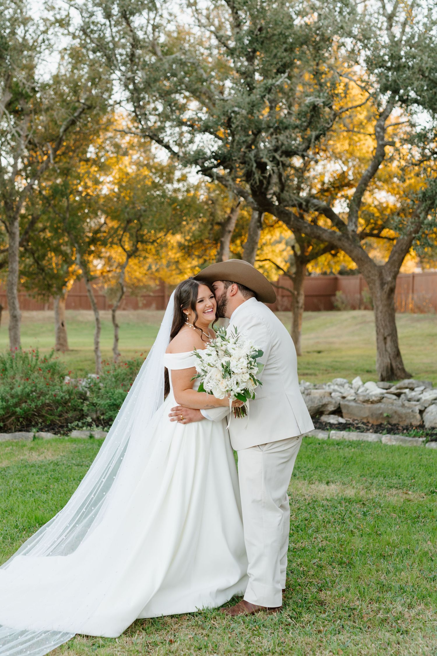 A wedding couple embraces outdoors in a garden setting with fall trees and golden sunlight in the background.