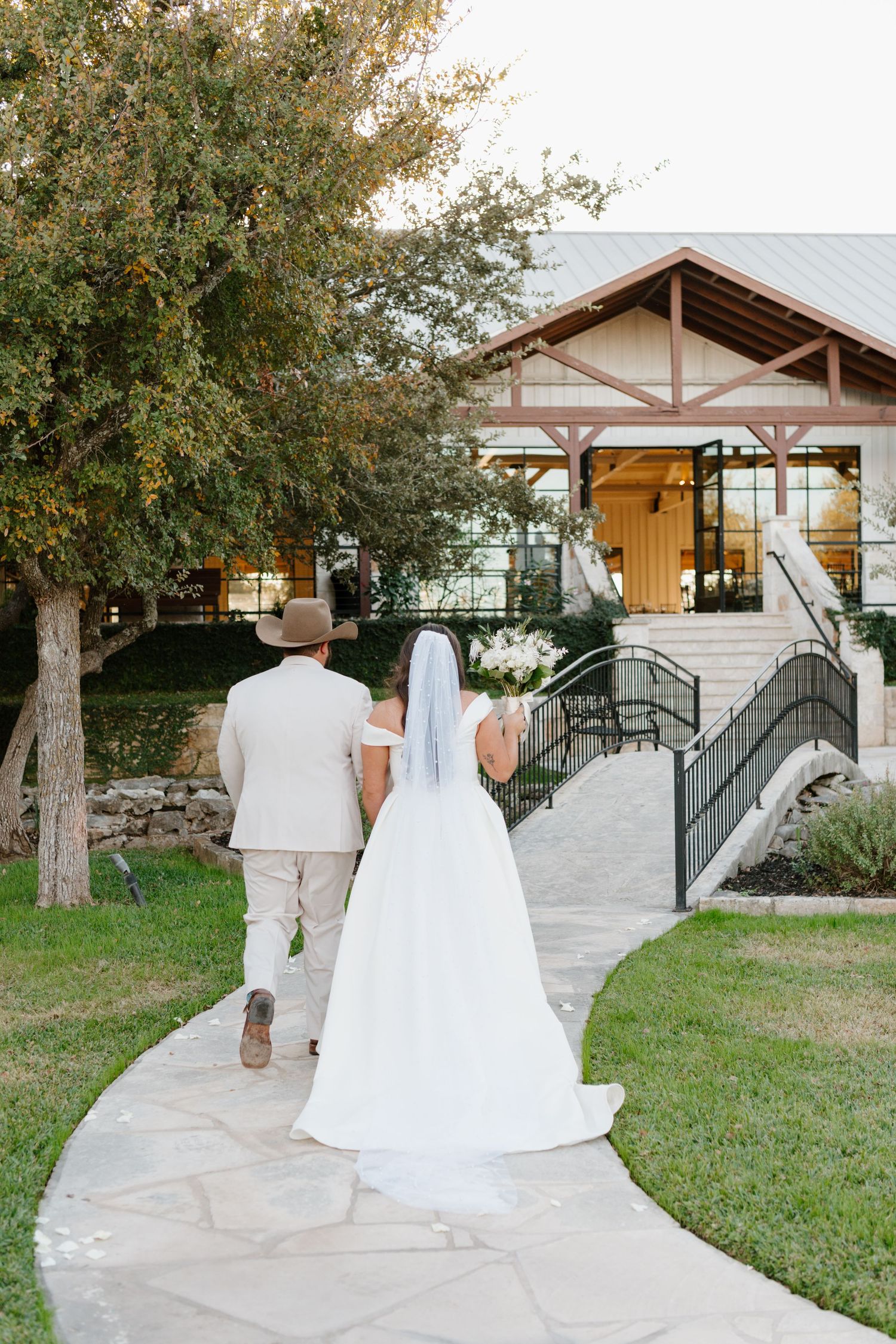 Couple walking up stone pathway towards rustic wedding venue entrance with wooden beams and white exterior.