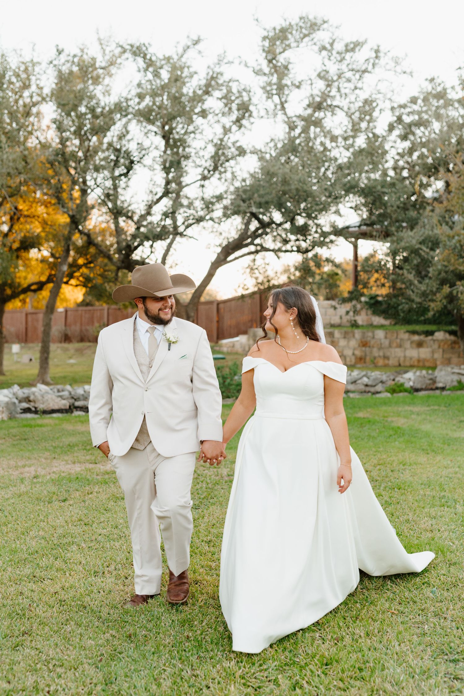 A couple in wedding attire walks hand in hand on grass with trees in the background during golden hour.