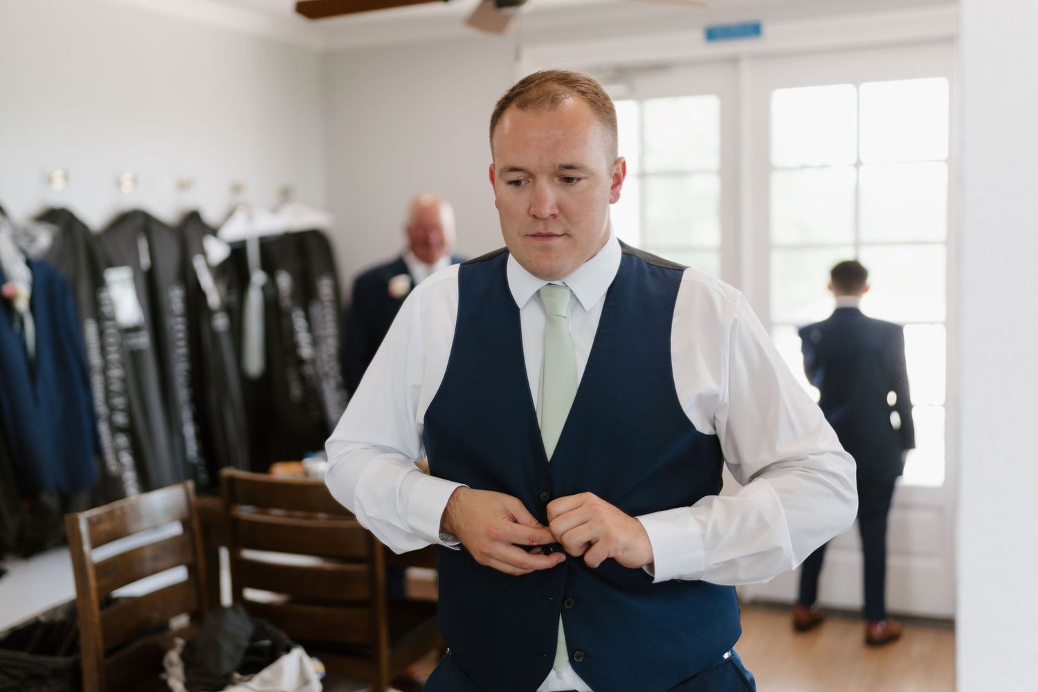 Groomsmen in navy suits and vests getting ready in a light-filled room.