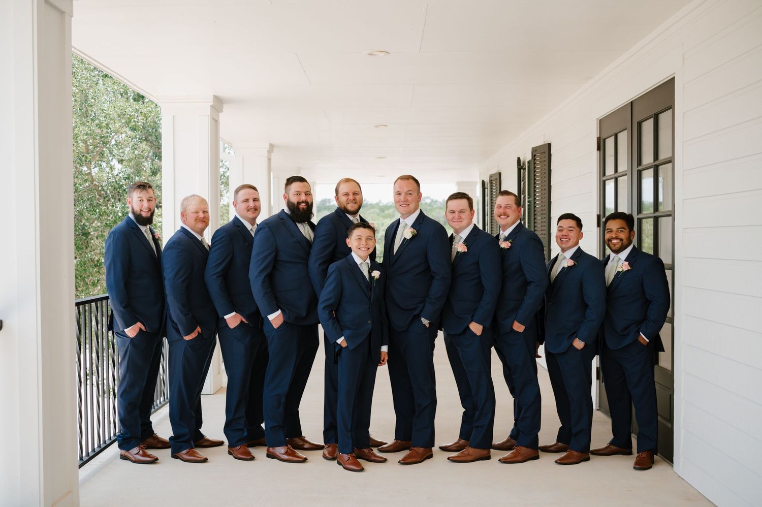 Groomsmen in navy suits lined up along a white columned porch for formal wedding photos.