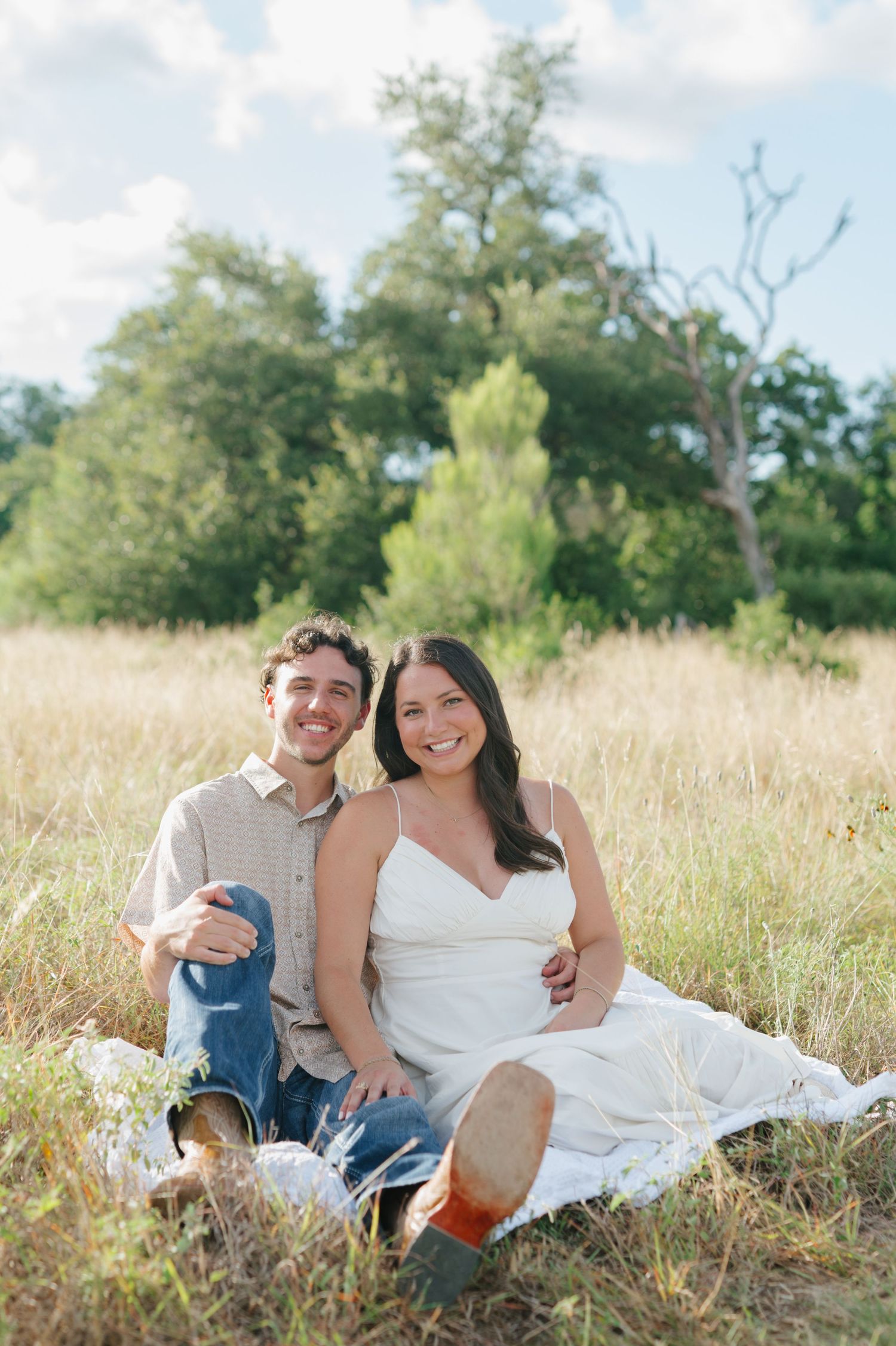 A couple relaxing together on a blanket in a sunlit meadow with trees in the background.