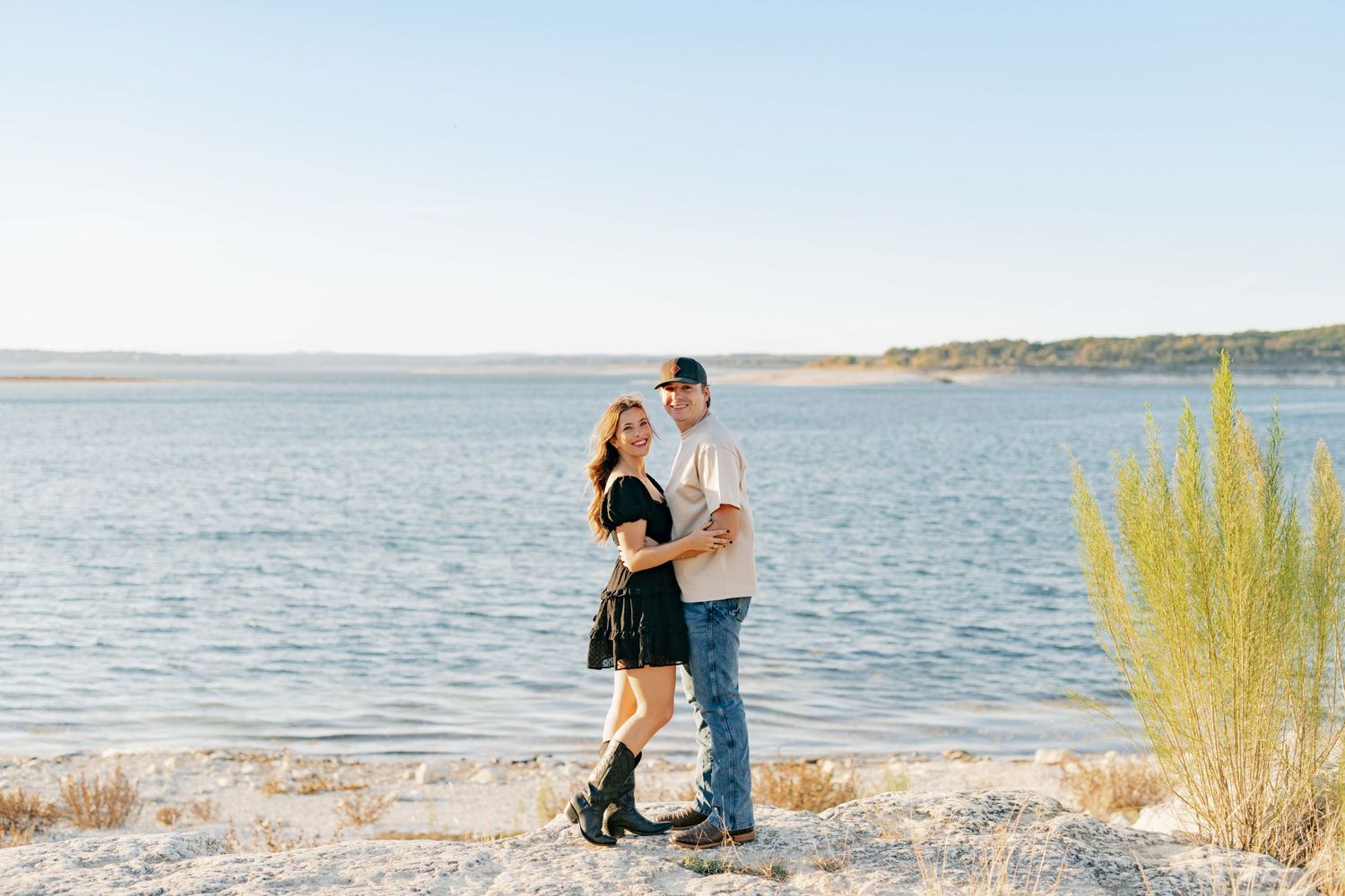 Couple sharing an intimate moment by the waterfront with tall grass and clear blue skies in the background.