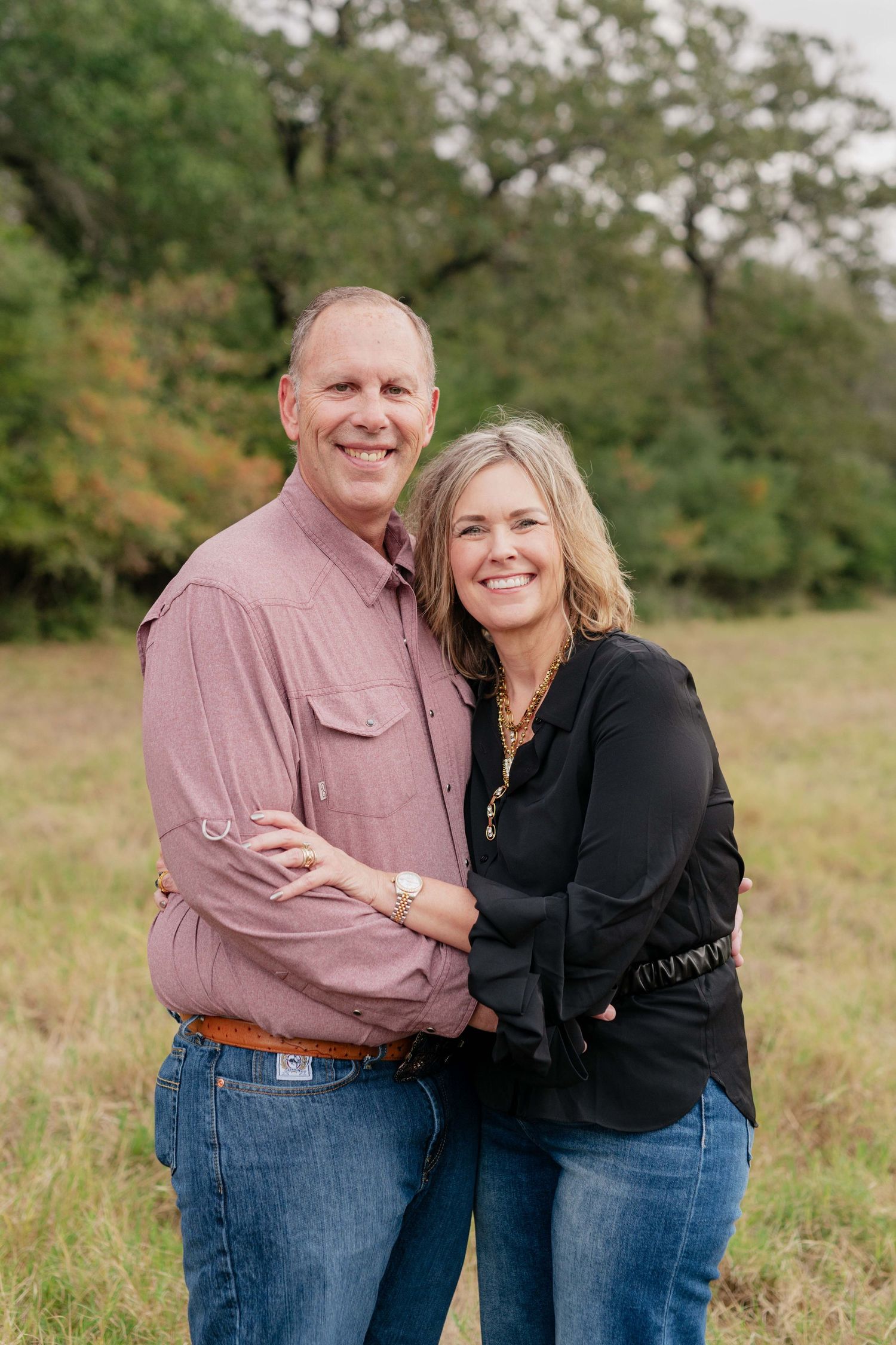 A couple embraces while smiling in a field with autumn foliage in the background.
