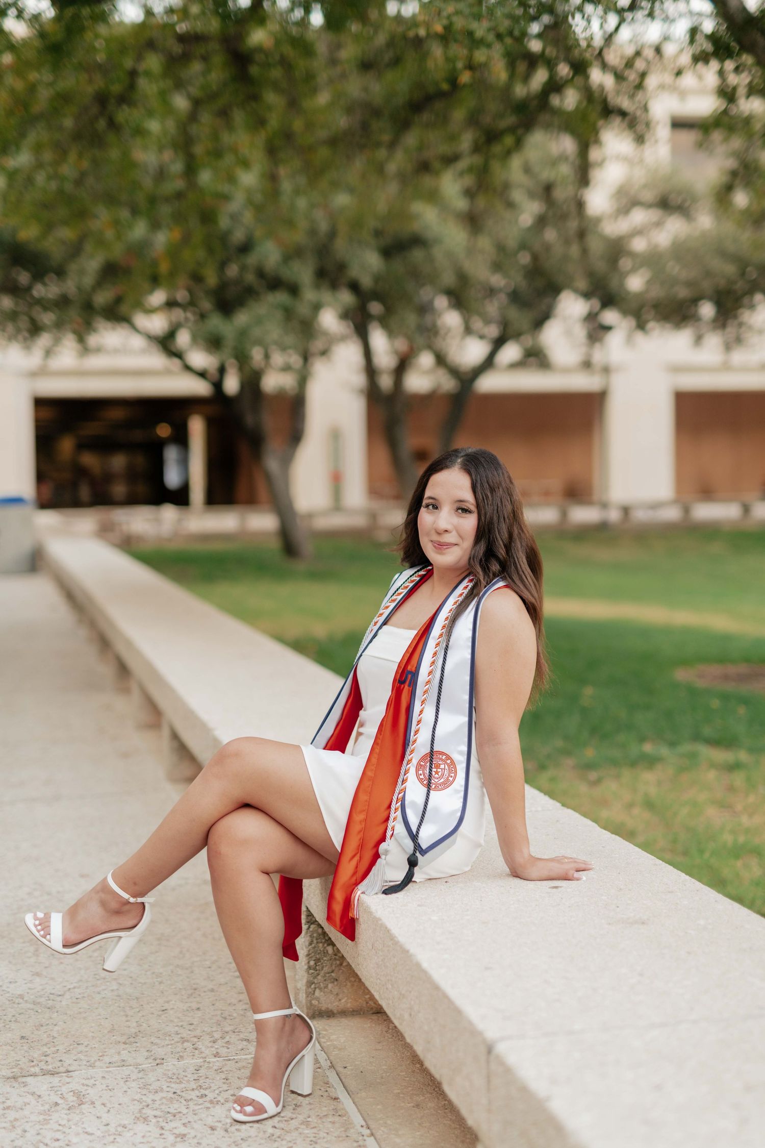 A graduate in white poses confidently on a concrete ledge near campus buildings.
