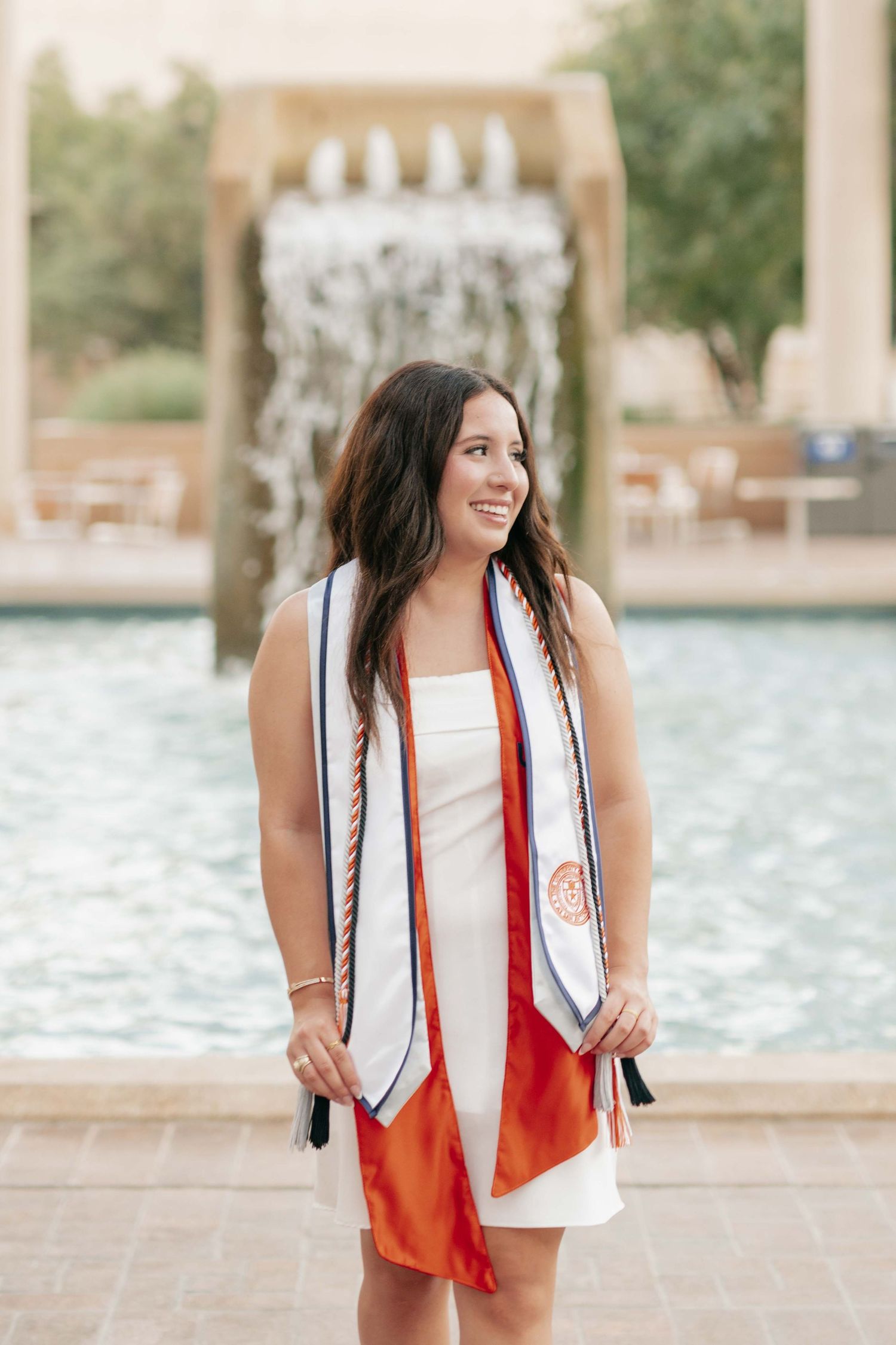 Graduate wearing white dress and graduation stole smiles by water feature.