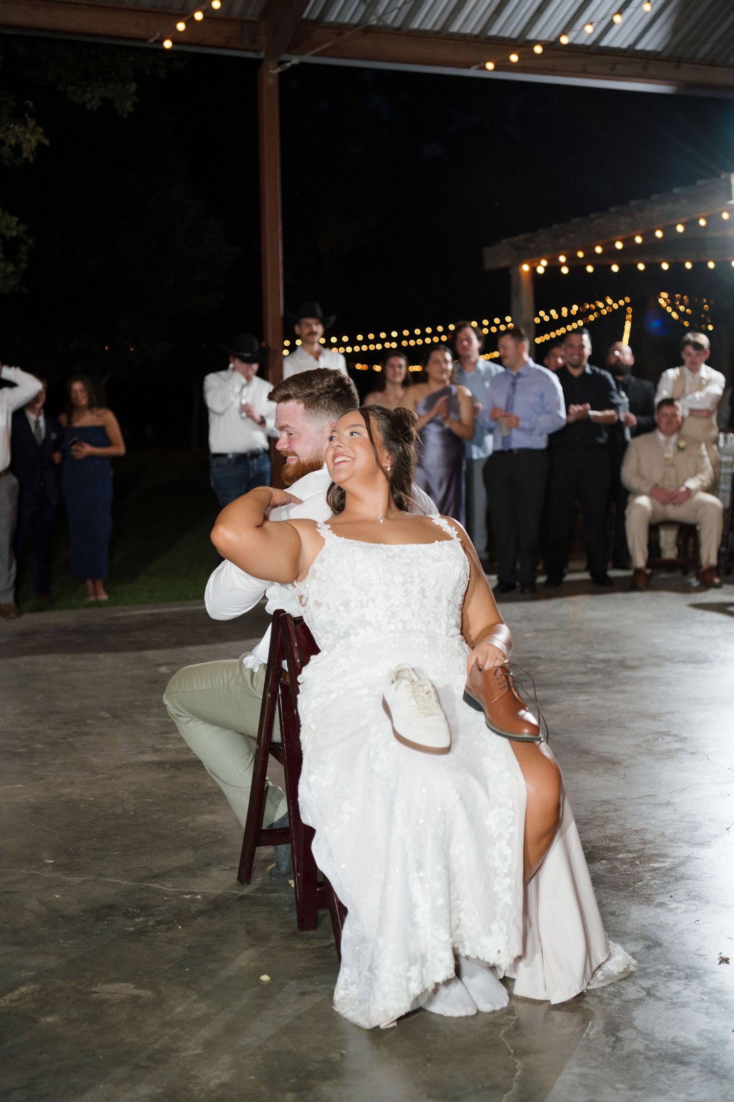 Bride sits smiling as groom removes her garter during outdoor evening wedding reception.