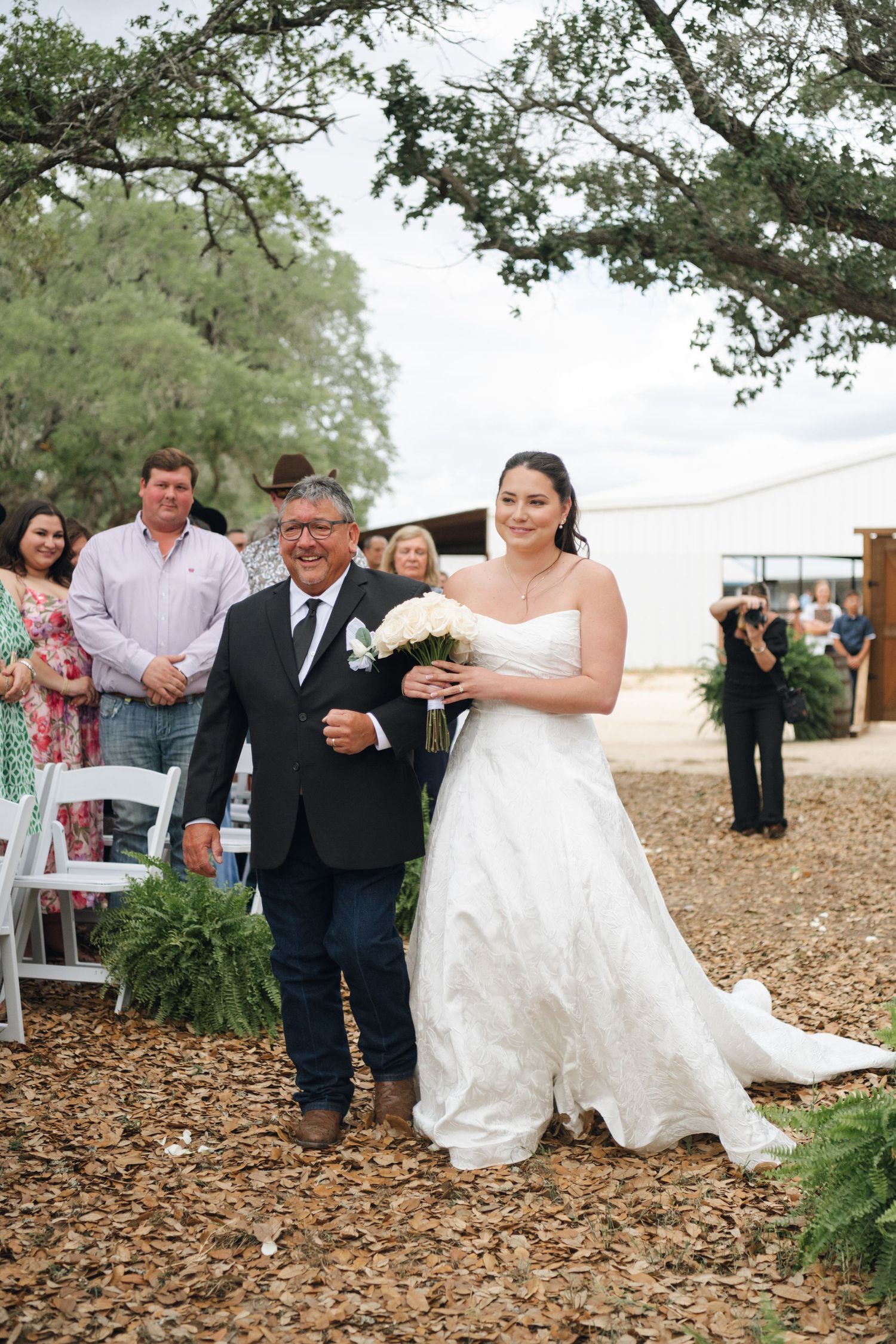 A smiling bride in a white strapless gown walks down the aisle with her father at an outdoor rustic wedding ceremony.