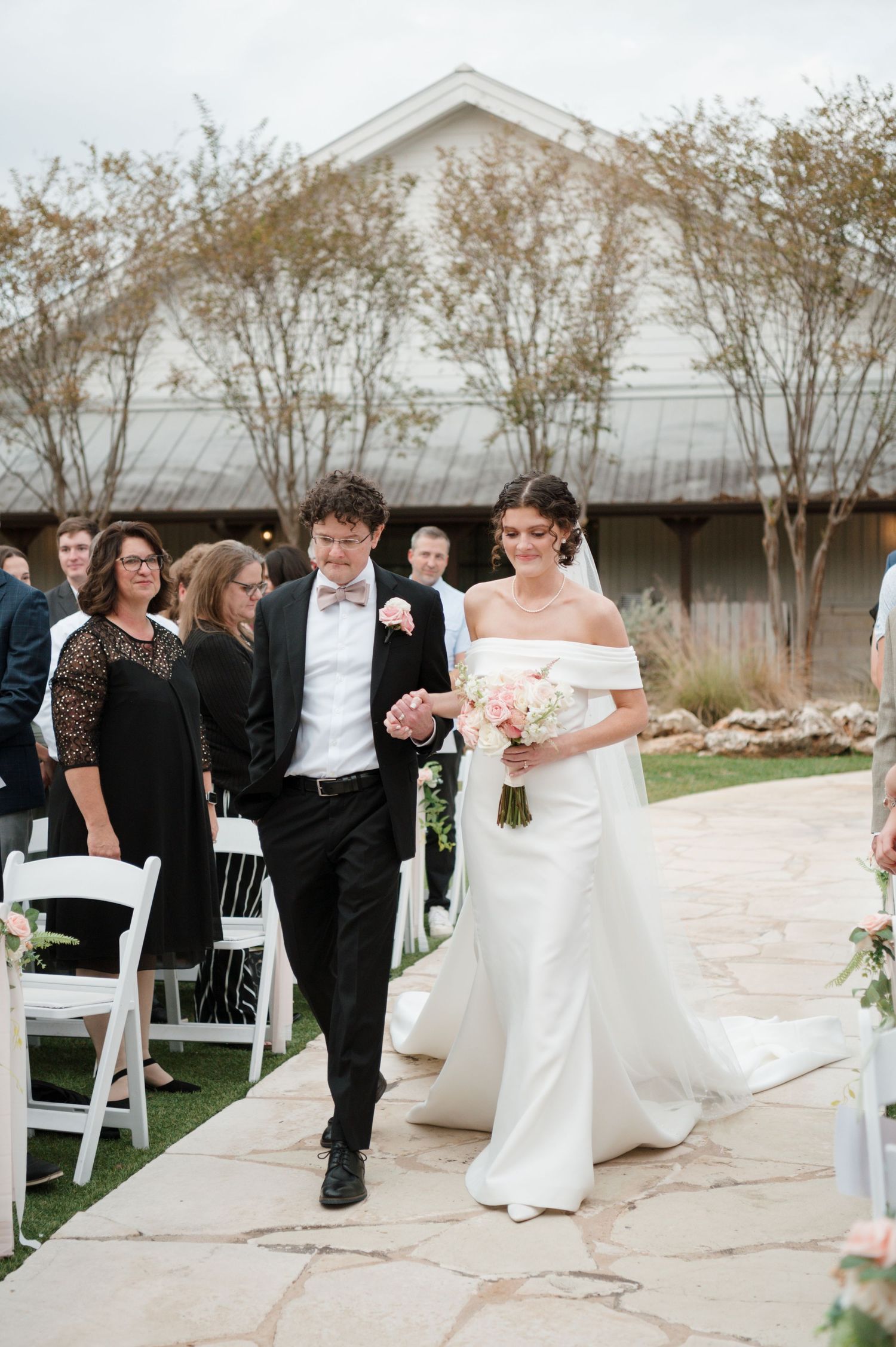 Wedding ceremony processional on an outdoor stone patio with white chairs and floral arrangements.