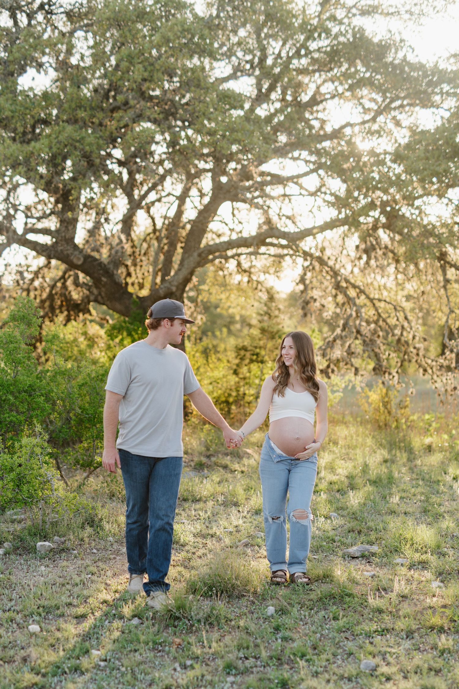 Couple holding hands in a grassy field beneath a large oak tree at sunset during a maternity photoshoot.