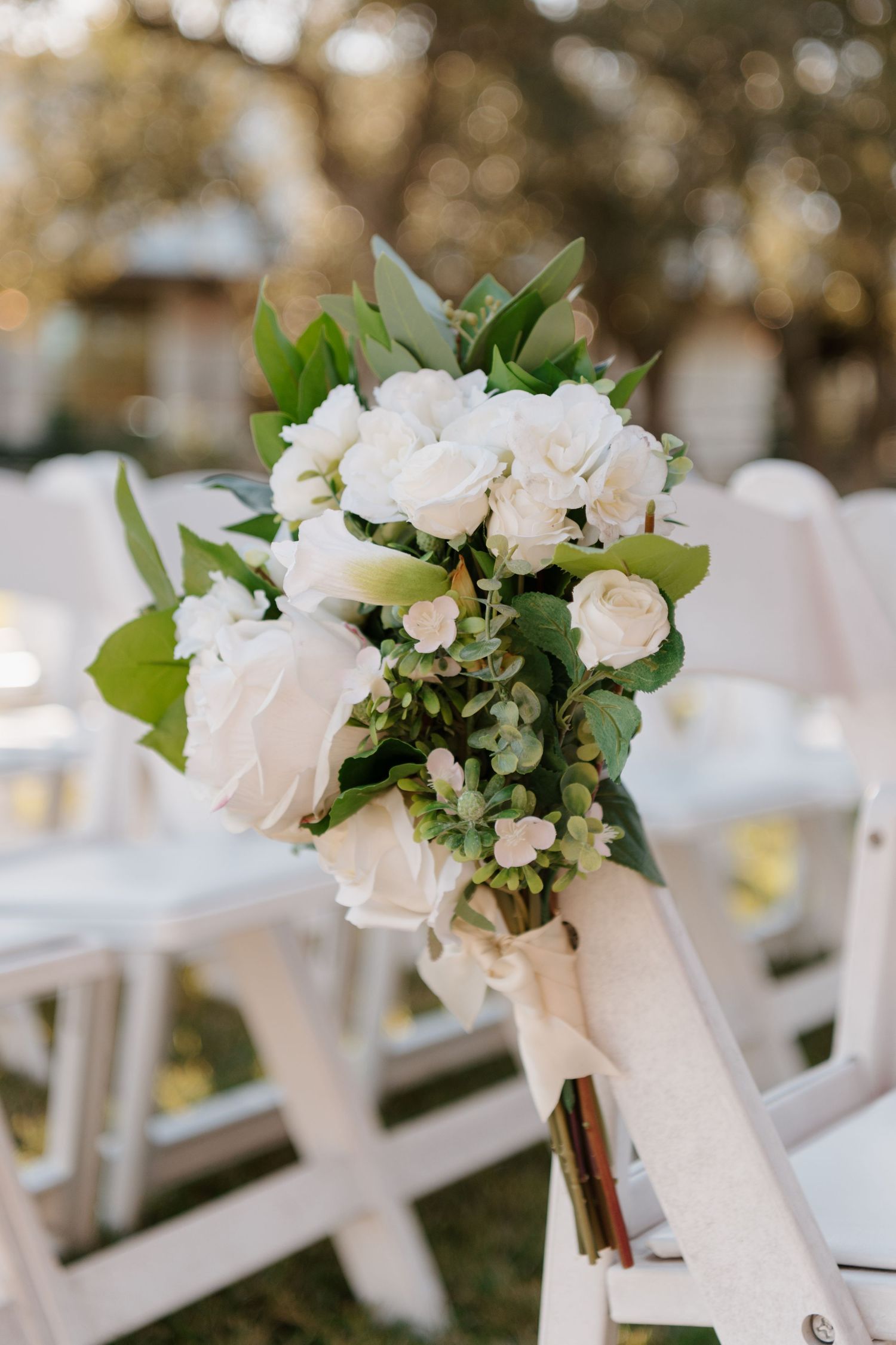 A delicate white rose bouquet with green foliage tied to a white ceremony chair for outdoor wedding decor.