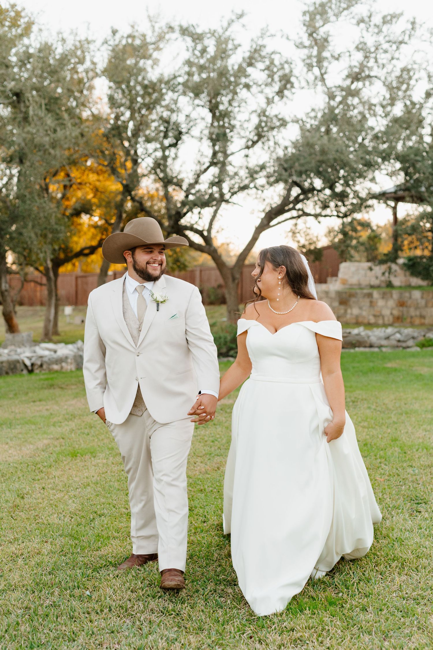 Couple in wedding attire walks hand in hand on grass with autumn trees and stone wall in background.