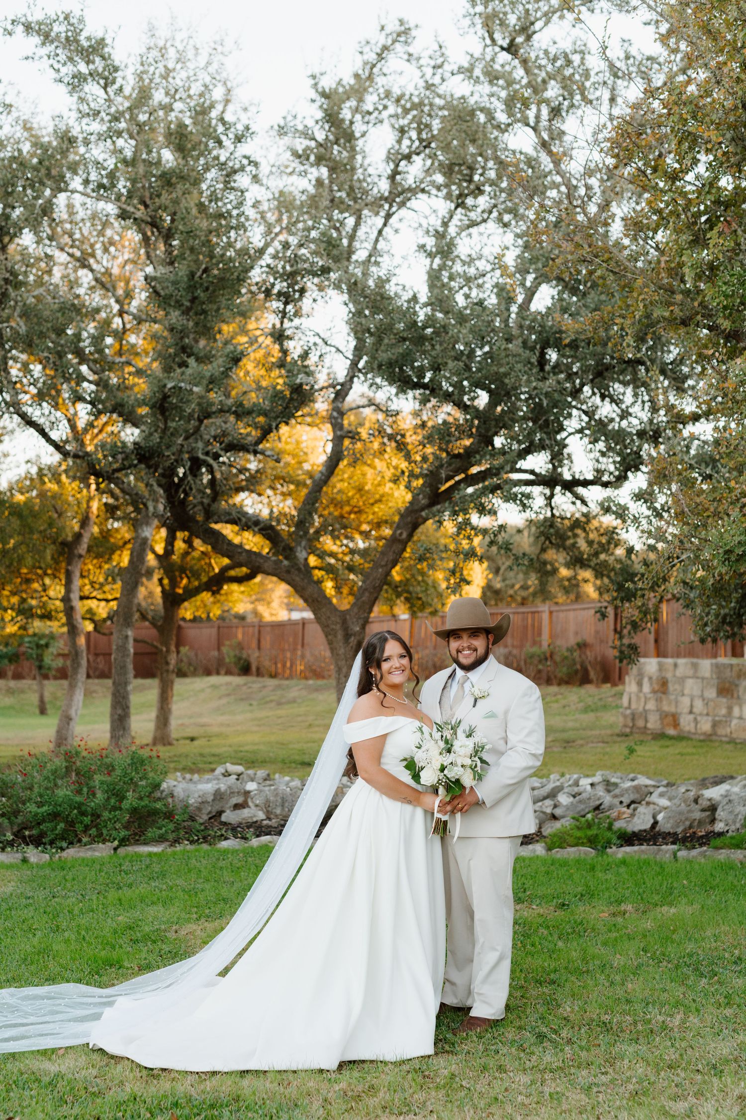 A romantic outdoor wedding scene with people posing under oak trees at golden hour, holding a white flower bouquet.