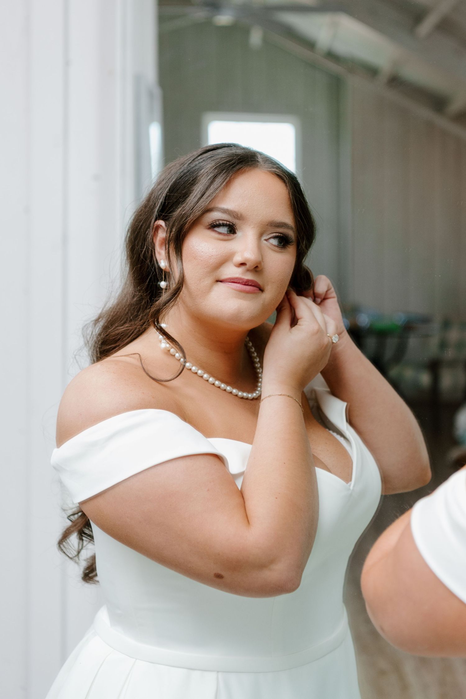 Someone in an elegant white off-shoulder dress and pearl necklace adjusts their earring while getting ready for a formal event.