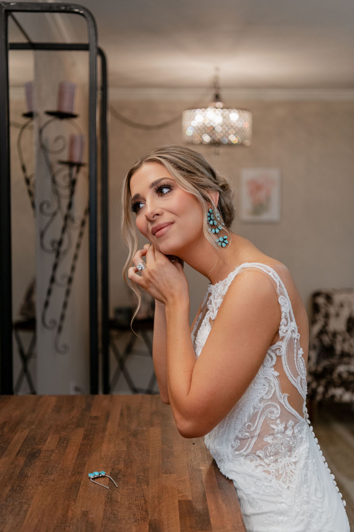 A bride in an elegant lace wedding dress with statement turquoise earrings sits at a wooden table beneath a crystal chandelier.