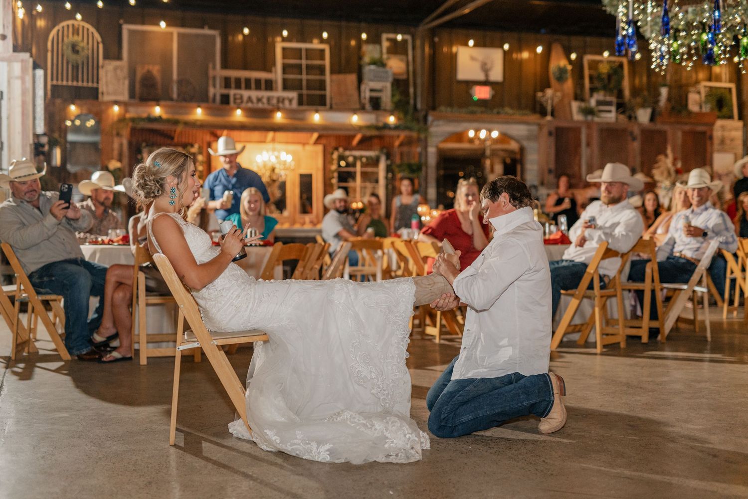A couple shares a playful moment during a wedding reception game in a rustic barn venue with twinkle lights overhead.