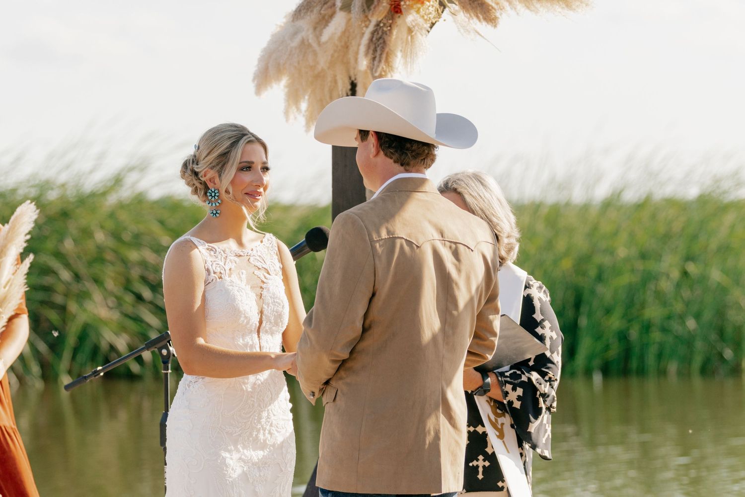A couple stands together during an outdoor wedding ceremony by a lake with tall reeds in the background.