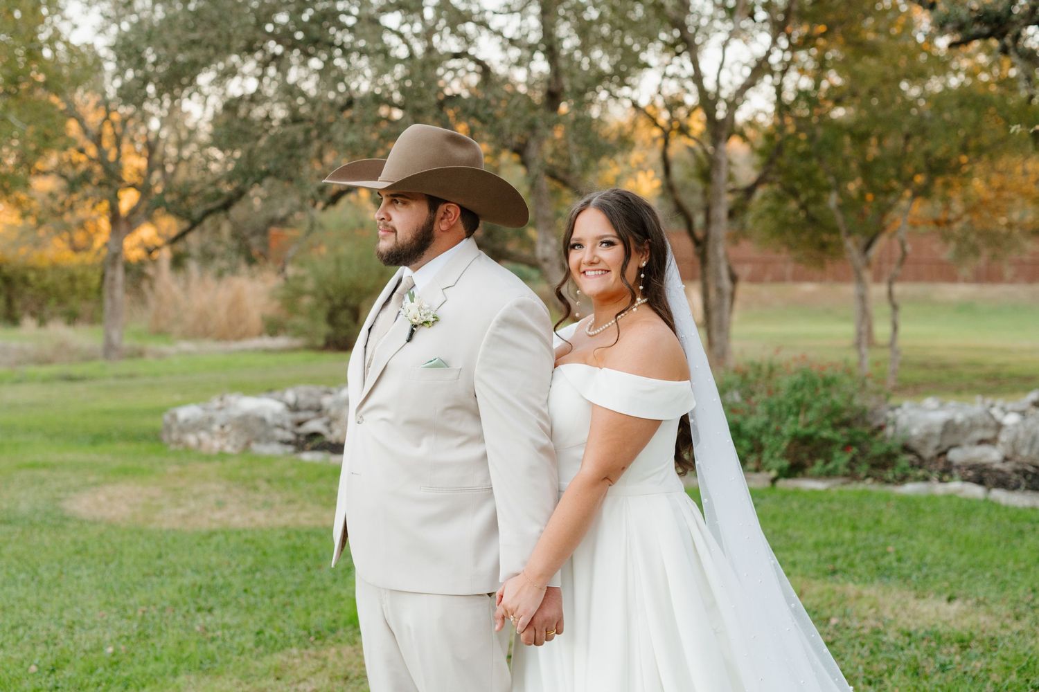 A bride and groom hold hands while standing back to back on a grassy lawn during golden hour at their outdoor wedding.