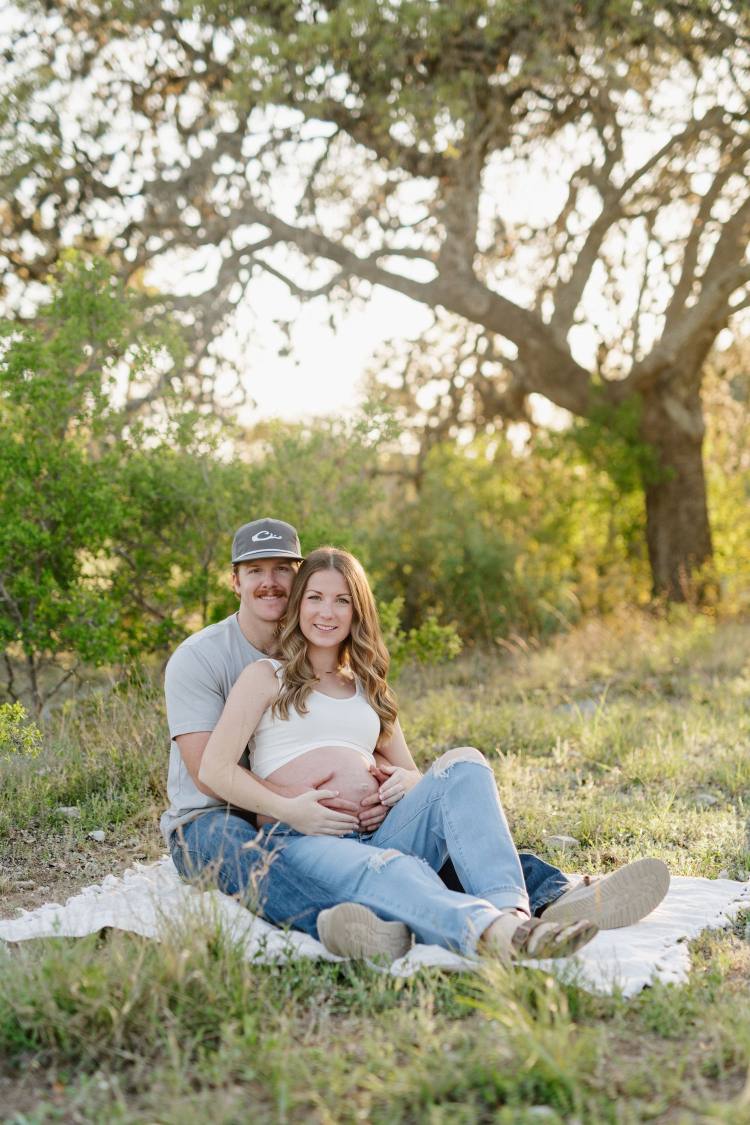 Couple sitting on blanket under tree during golden hour in outdoor maternity photo shoot.