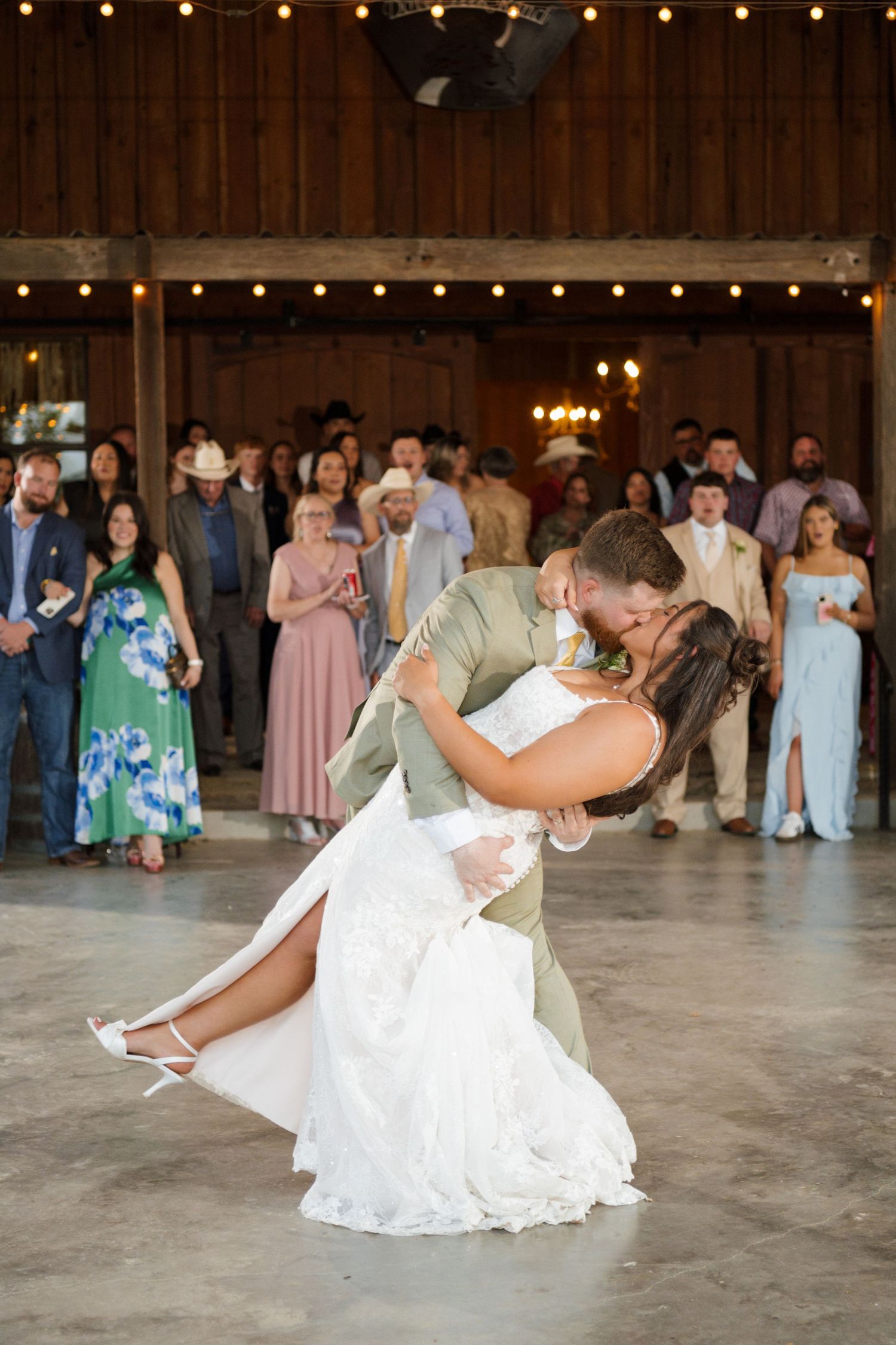 Groom dips bride for a romantic kiss during first dance in a barn reception with string lights.