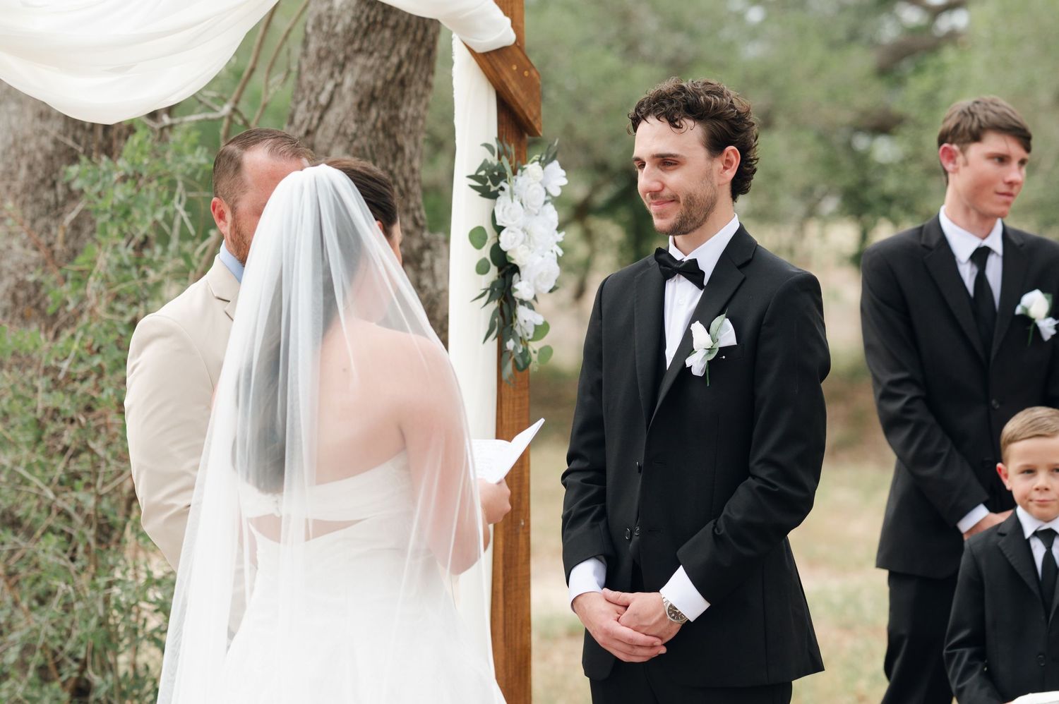 Groom in black tuxedo smiles at bride in white veil during outdoor wedding ceremony with floral arch.