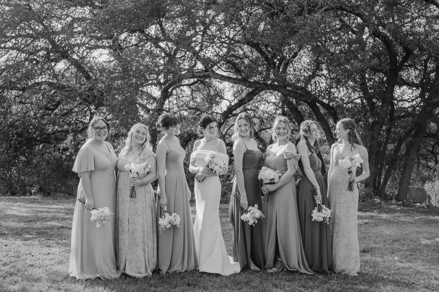 Wedding party in gray dresses and white bouquets poses together in a garden setting for formal portraits.