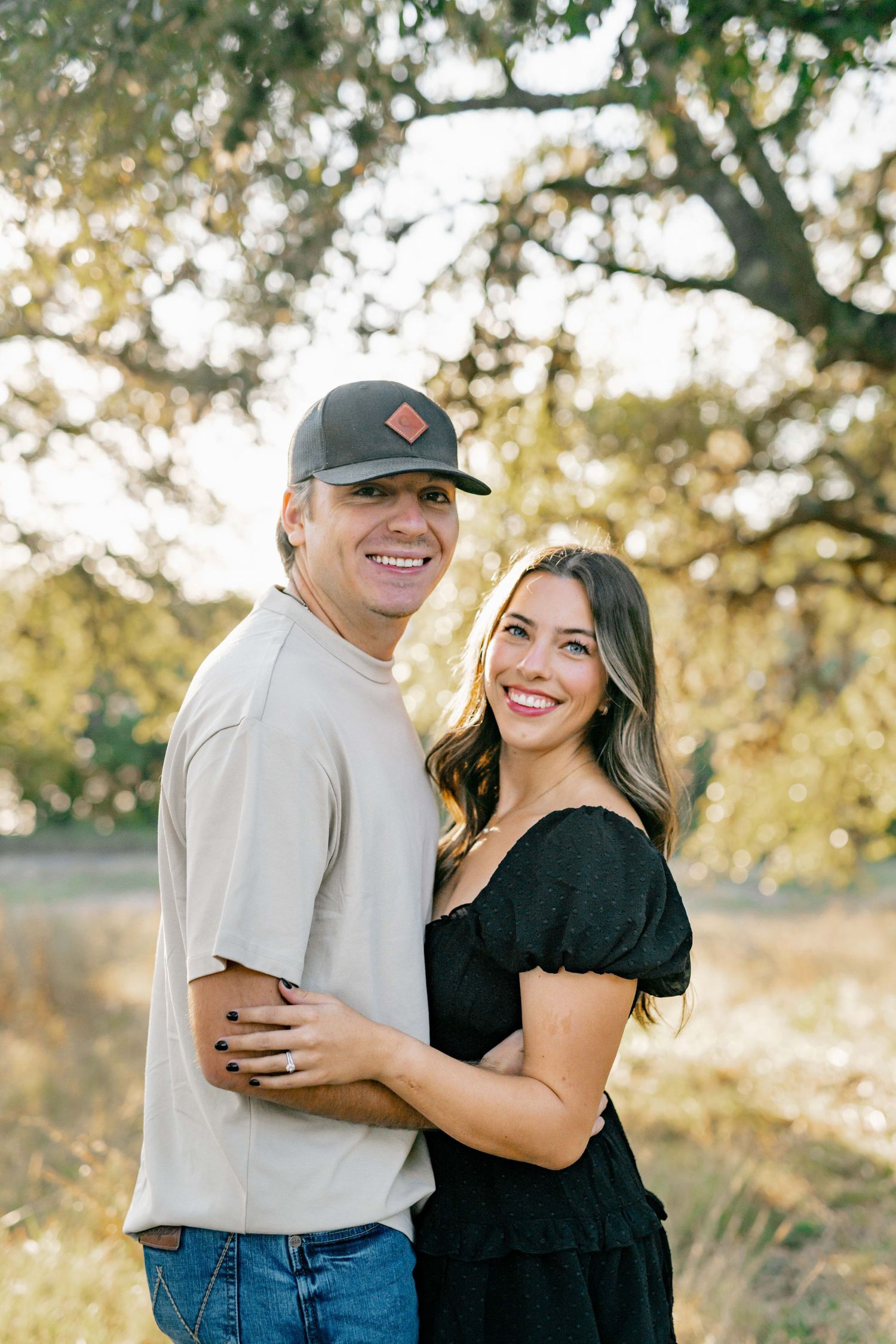 A couple embraces while posing together outdoors with golden sunlit trees in the background.