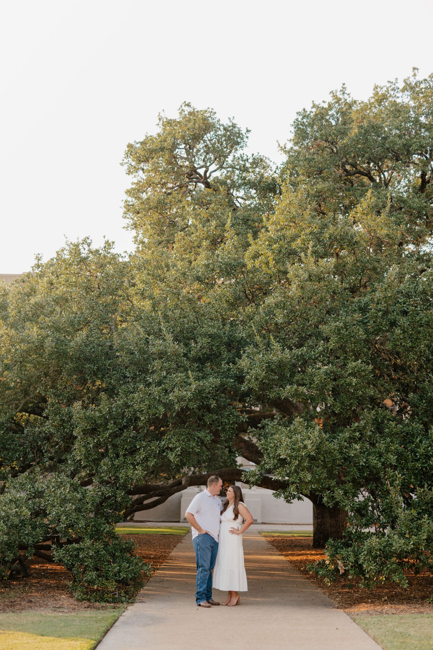 A couple stands together on a pathway beneath towering green trees.