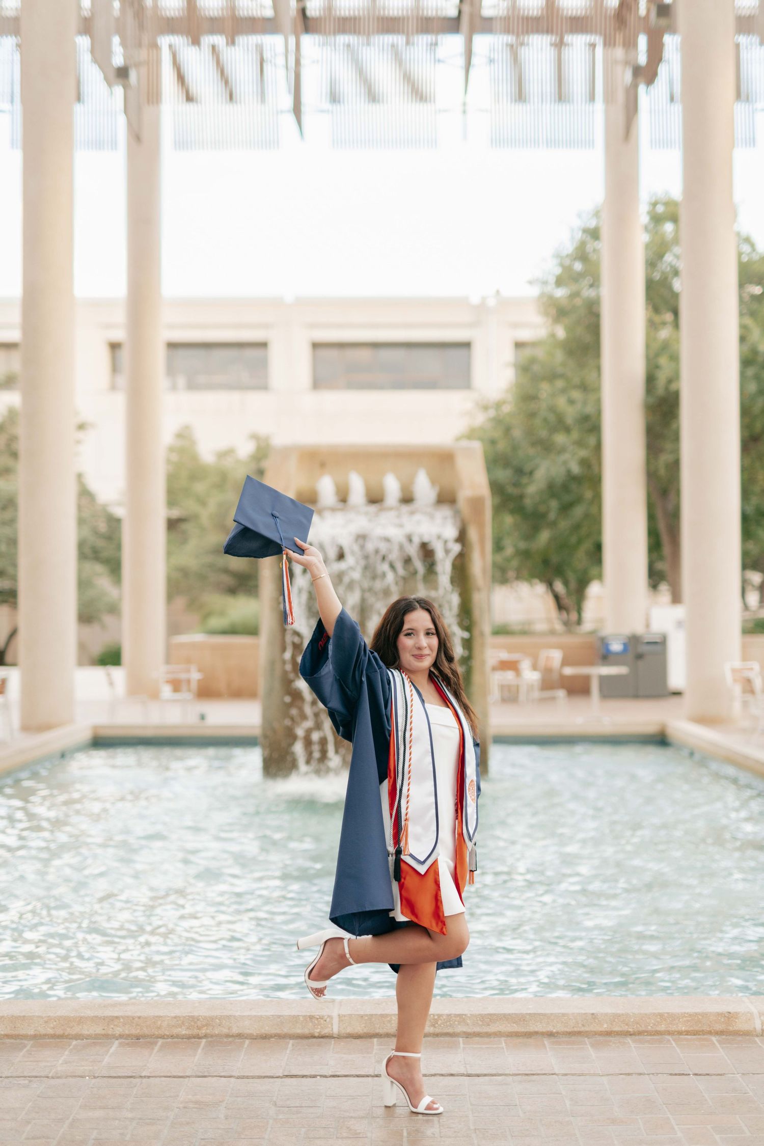 Graduate celebrates by fountain wearing graduation gown and throwing cap.