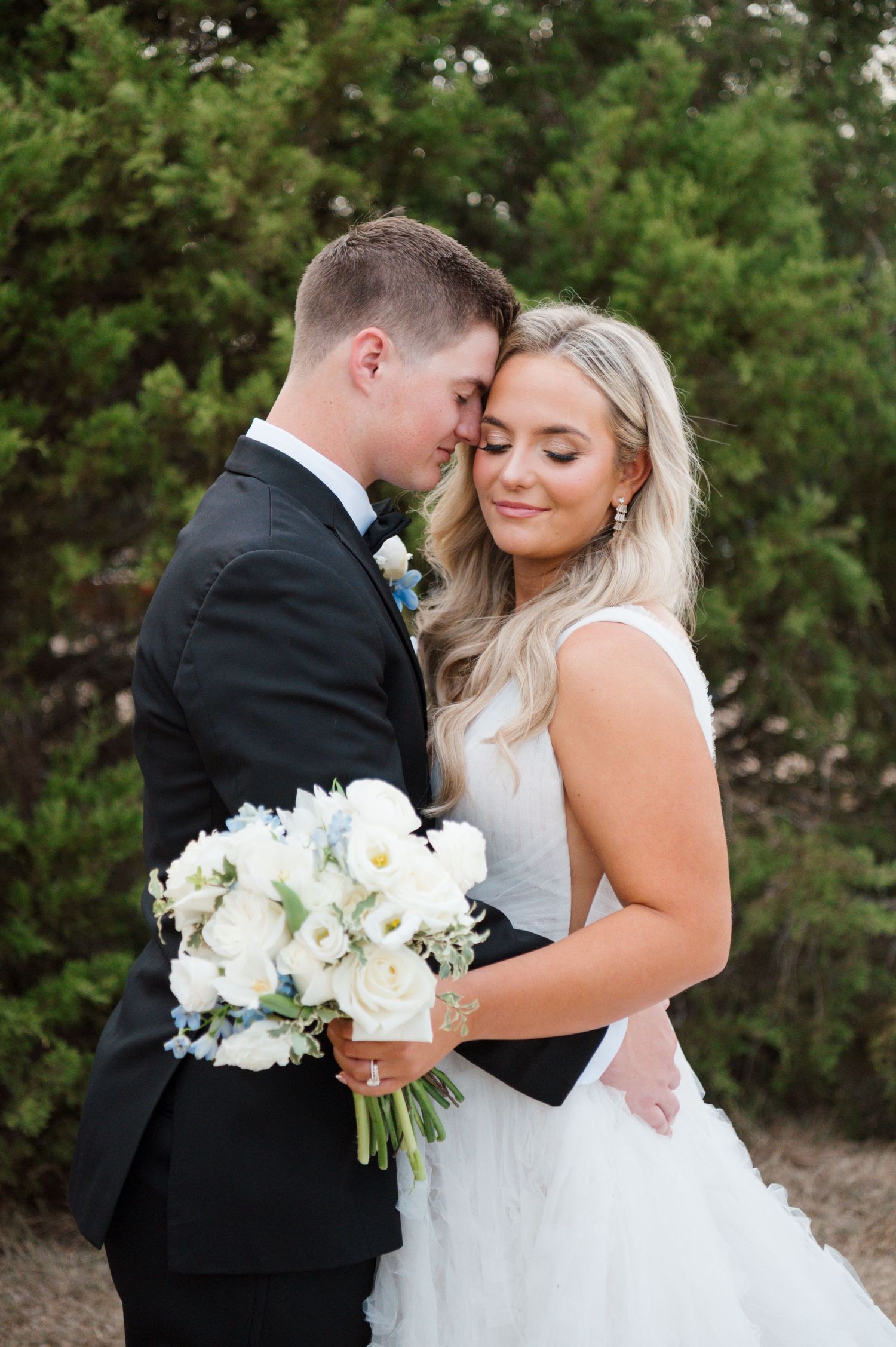 A bride and groom embrace each other in a scenic outdoor setting with lush greenery in the background.
