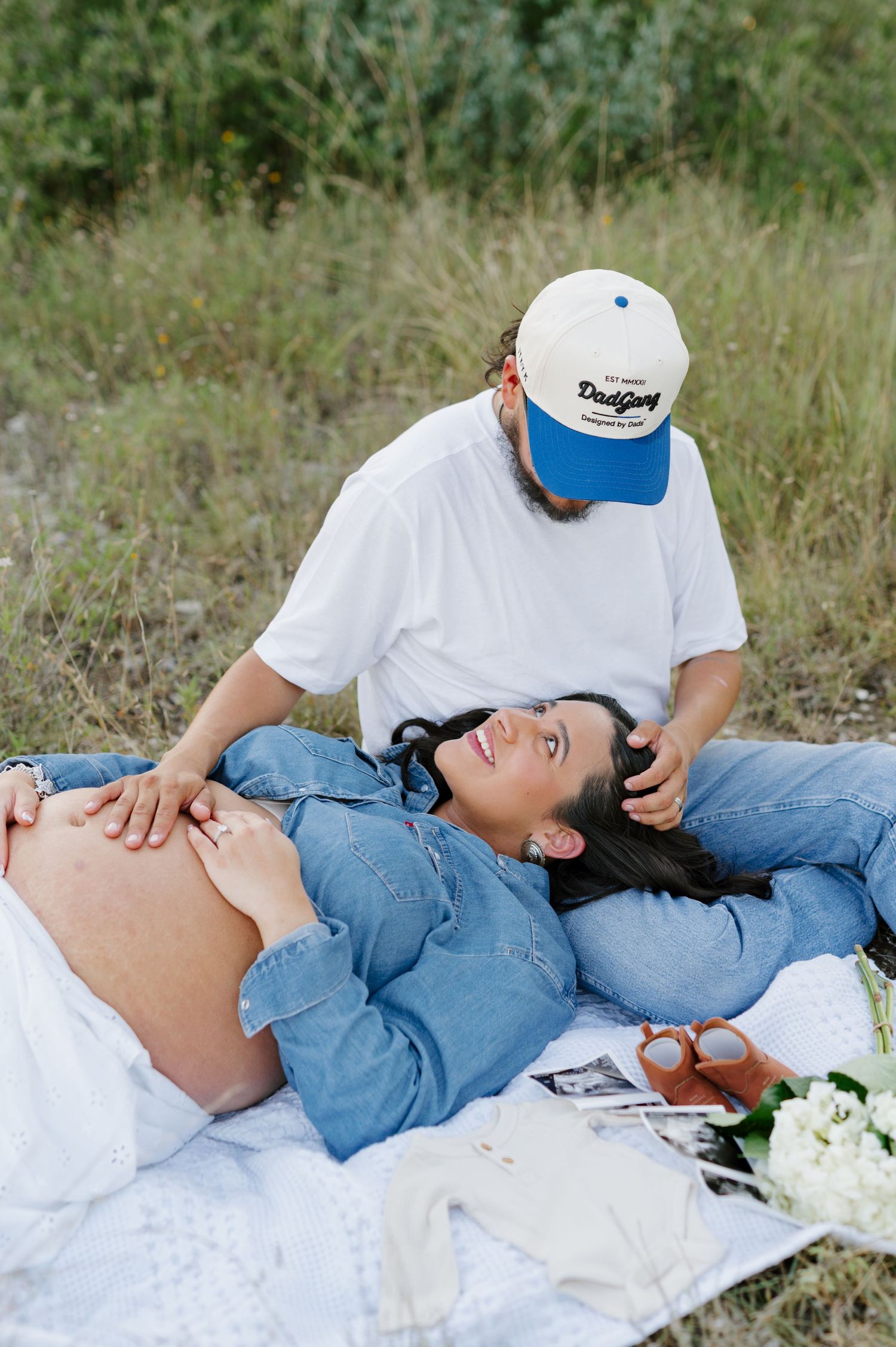 Couple relaxes on white blanket during outdoor maternity photoshoot with flowers nearby.