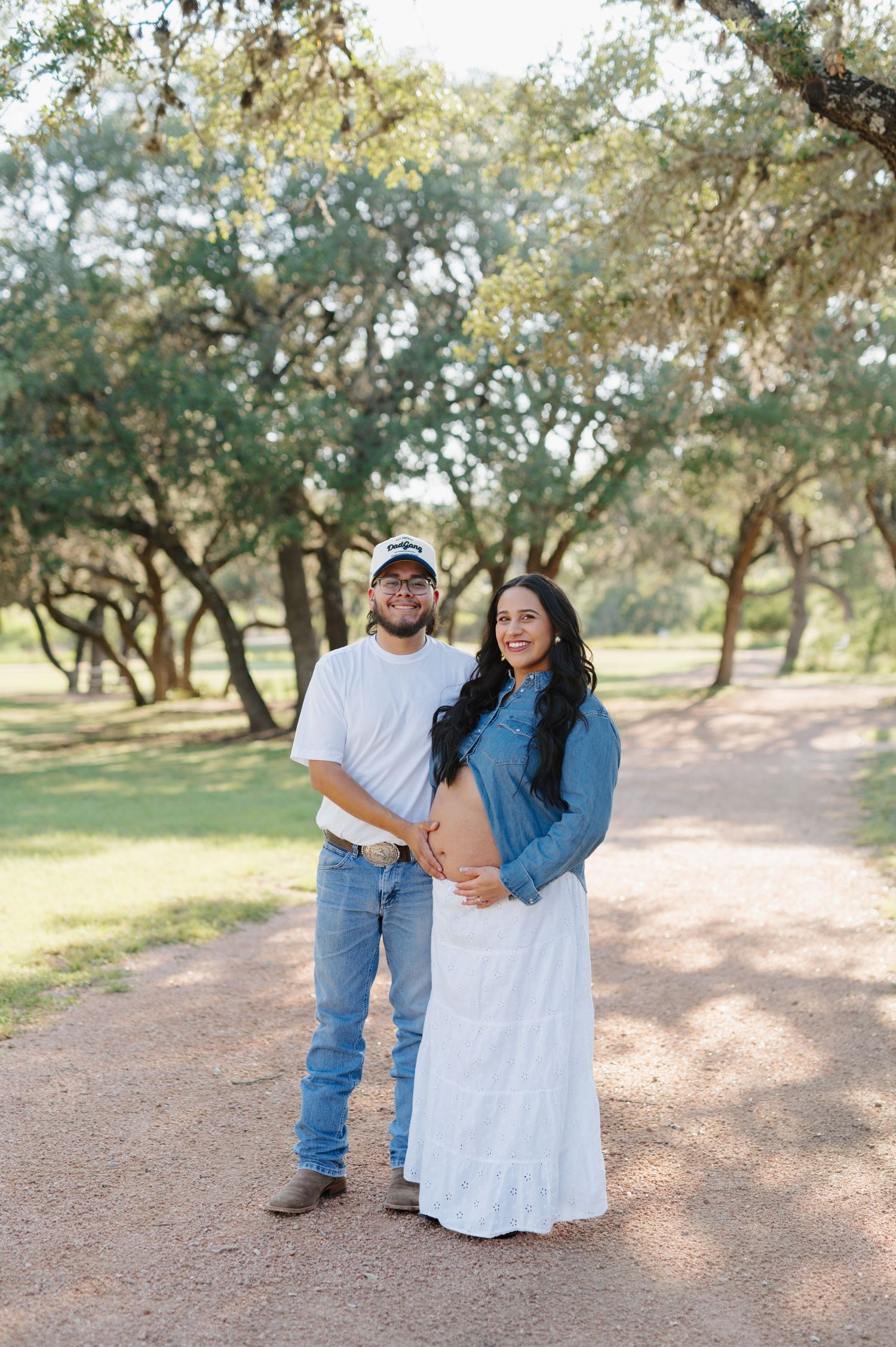 A couple poses together on a dirt path surrounded by oak trees in a romantic maternity photoshoot.